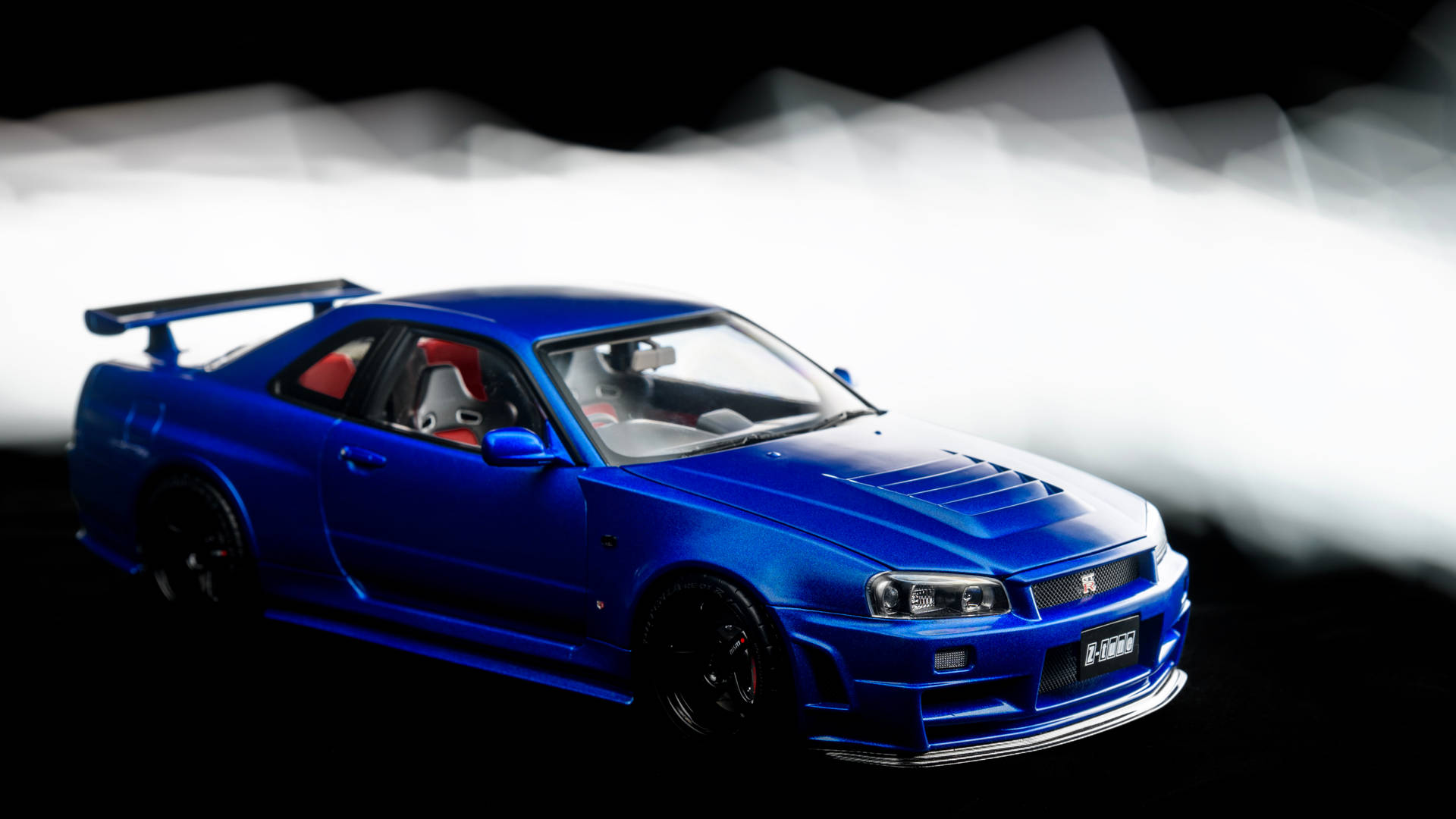 藍色日產Skyline GT-R模型車，配有紅色座椅和黑色車輪，在煙霧繚繞的攝影棚燈光下。 — 日產 Skyline GTR R34 壁紙