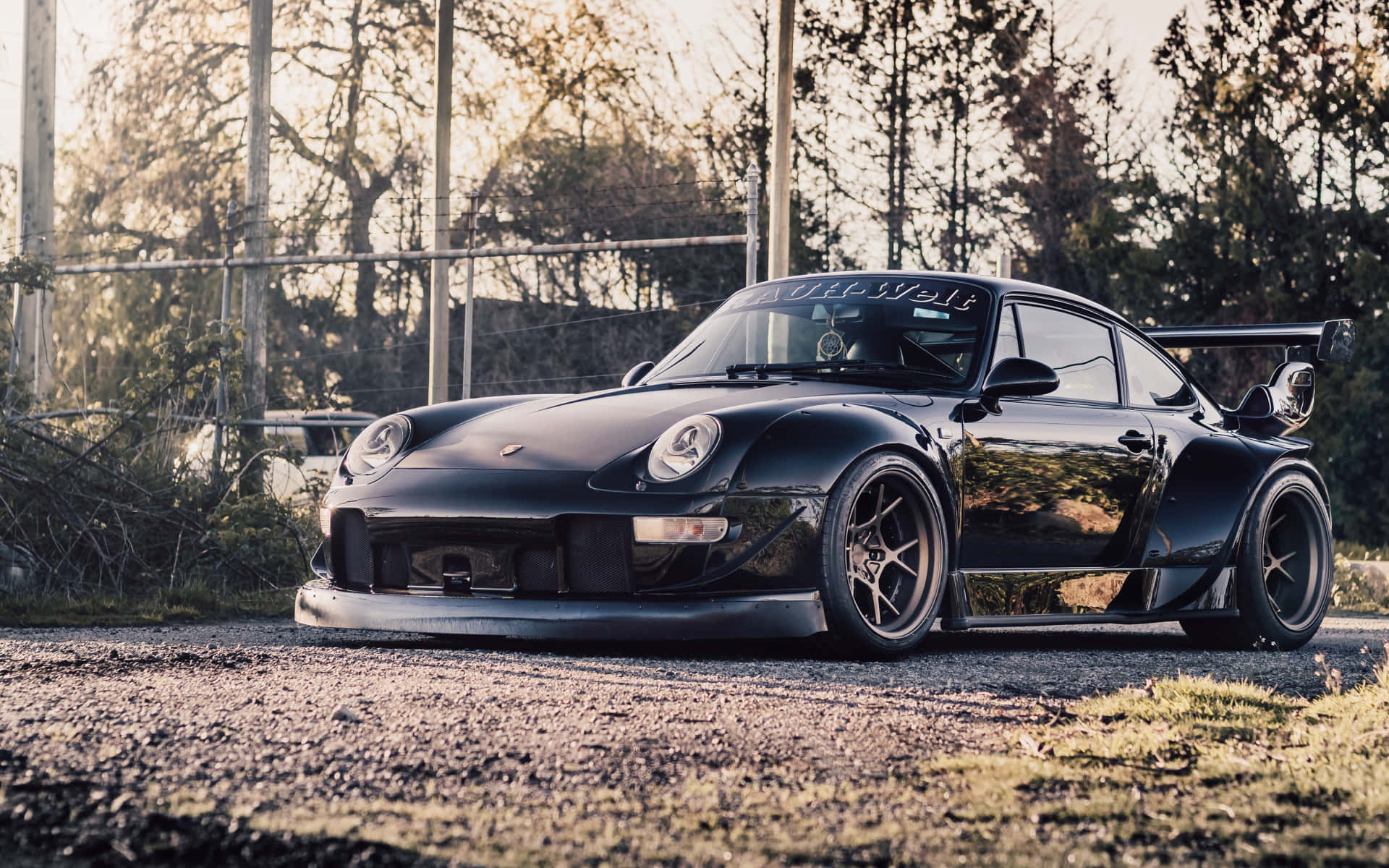  壁紙 — 一輛亮黑色保時捷911 GT1跑車，配備寬體車身套件和後尾翼，在黃金時段停泊在樹木附近的碎石路上。