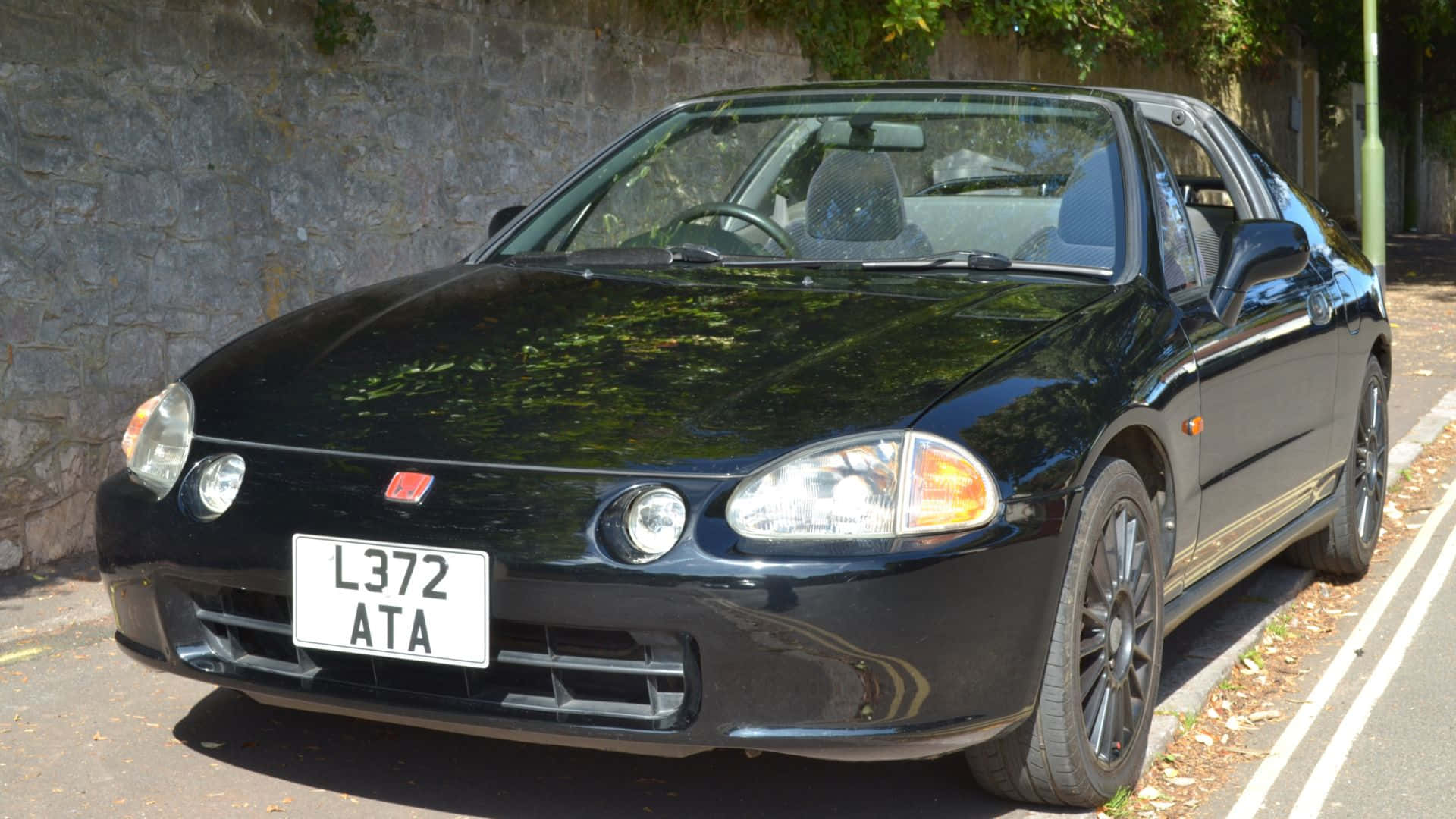 Honda Civic Del Sol 桌布 壁紙 — 一輛亮黑色本田Civic Type R停放在陽光明媚的街道上，旁邊是石牆，上方有綠色樹葉。