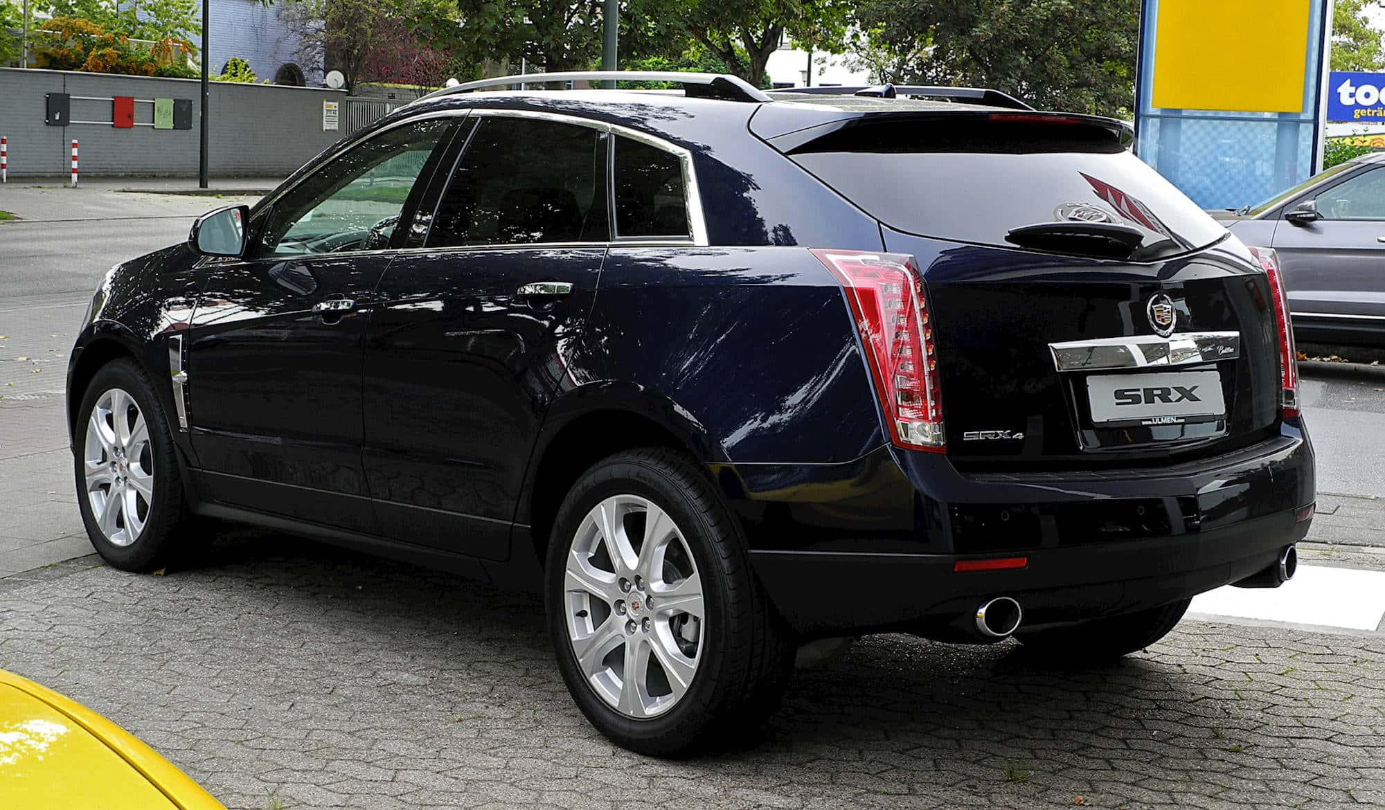 Cadillac Srx 桌布 壁紙 — 一輛亮黑色的Cadillac SRX SUV停泊在鵝卵石街道上，背景有樹木和黃色標誌。