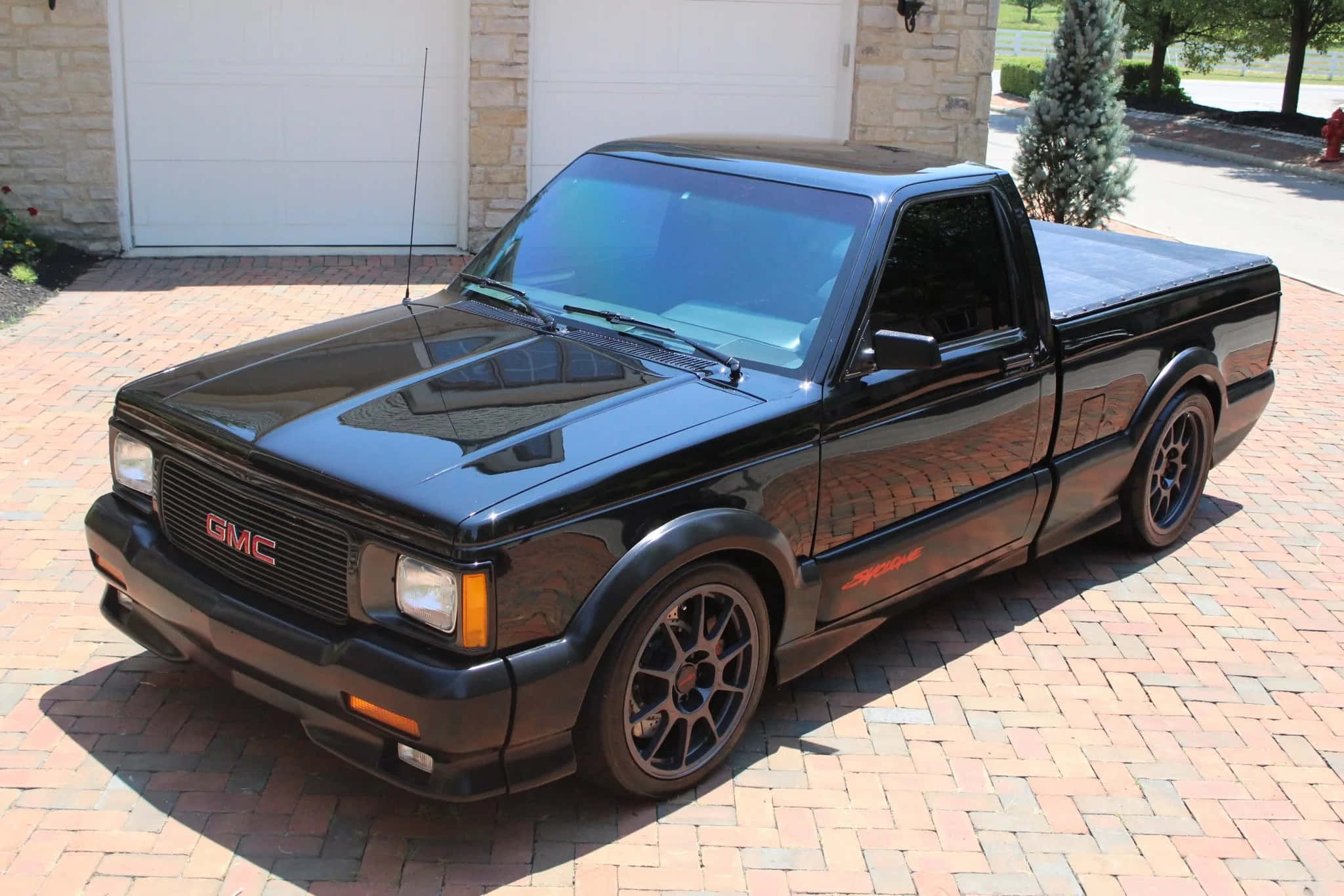Gmc Syclone 桌布 壁紙 — 一輛亮黑色的GMC Syclone皮卡車停在石車庫前的紅磚車道上。