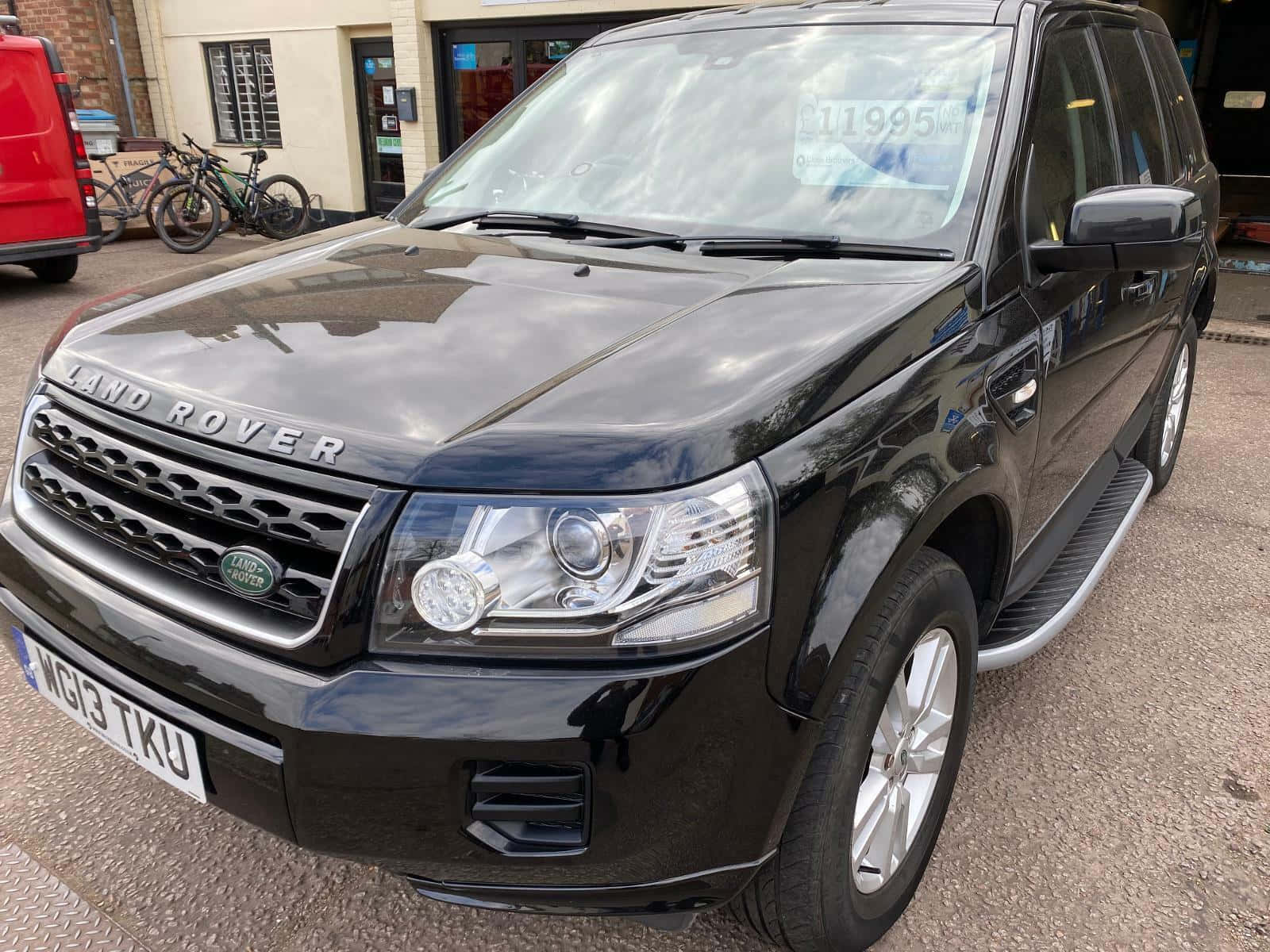 Land Rover Freelander 桌布 壁紙 — 一輛亮黑色的Land Rover Freelander停在鋪砌的停車場，背景有自行車和紅色廂型車。