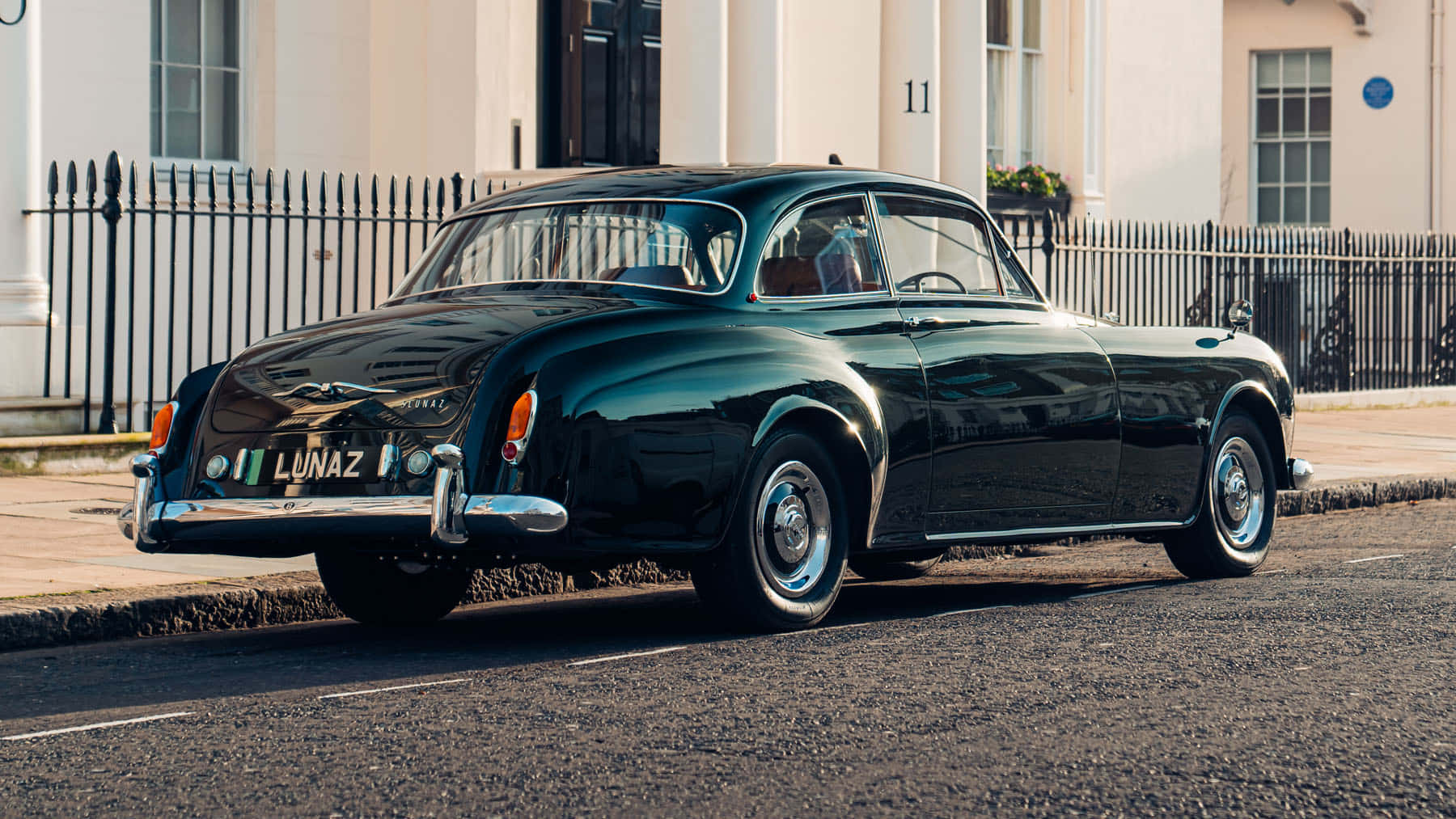 Bentley S2 桌布 壁紙 — 一輛亮黑色復古跑車停放在陽光明媚的城市街道上，旁邊是白色建築和黑色圍欄。