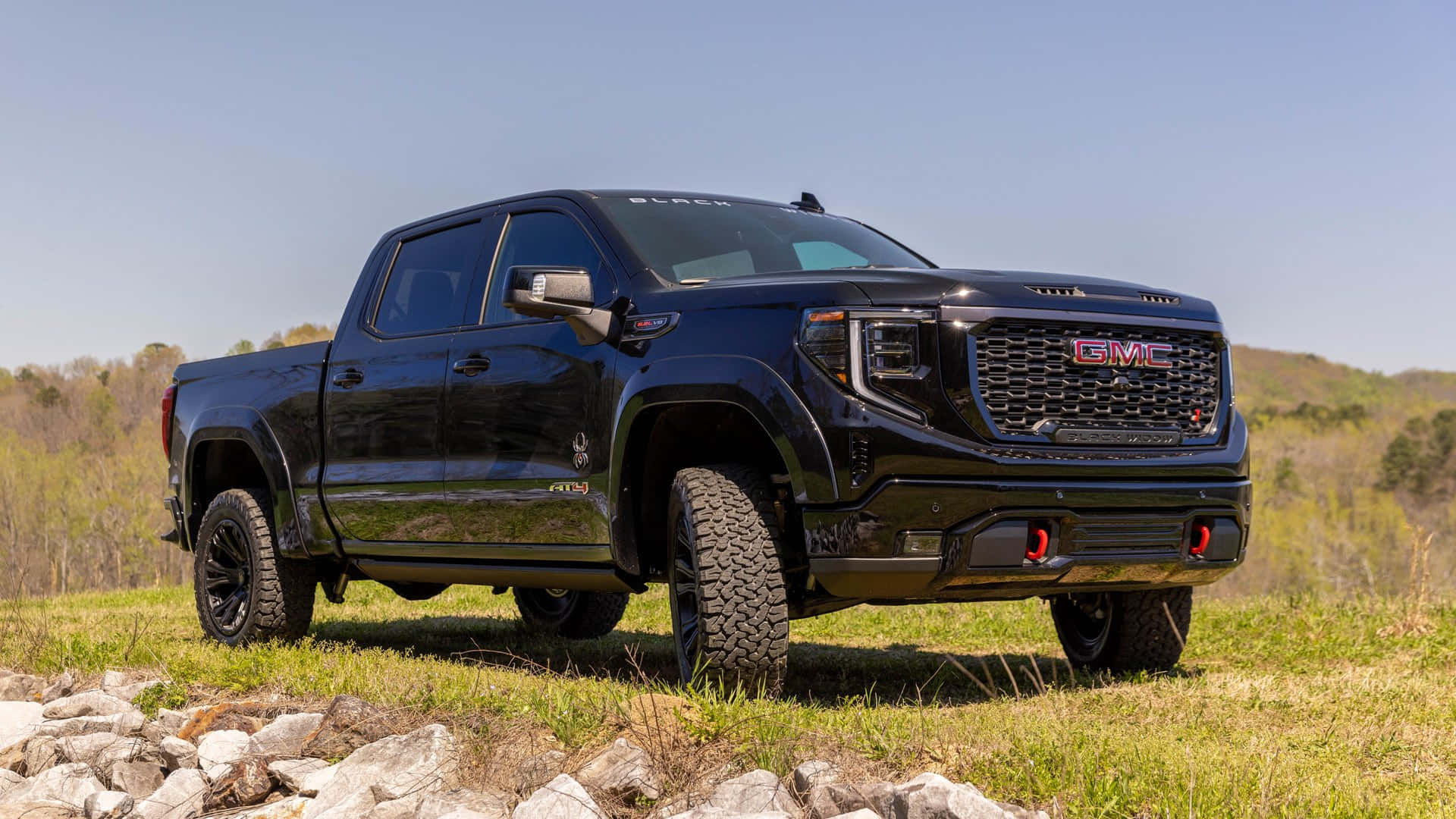 Gmc Sierra 桌布 壁紙 — 一輛亮黑色GMC Sierra AT4X pickup truck在晴朗藍天下，堅固地停放在有岩石和遠處樹木的草坡上。