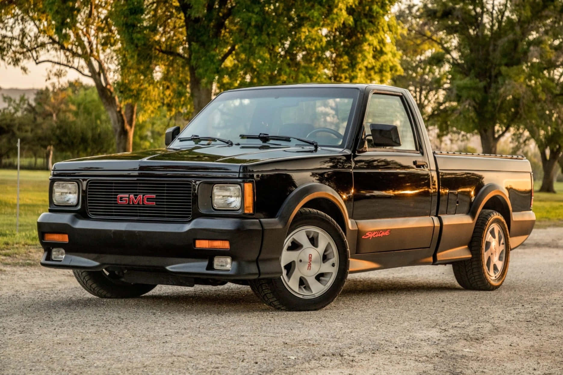 Gmc Syclone 桌布 壁紙 — 一輛亮黑色GMC Syclone皮卡車停在碎石上，樹木在金色夕陽下閃耀。