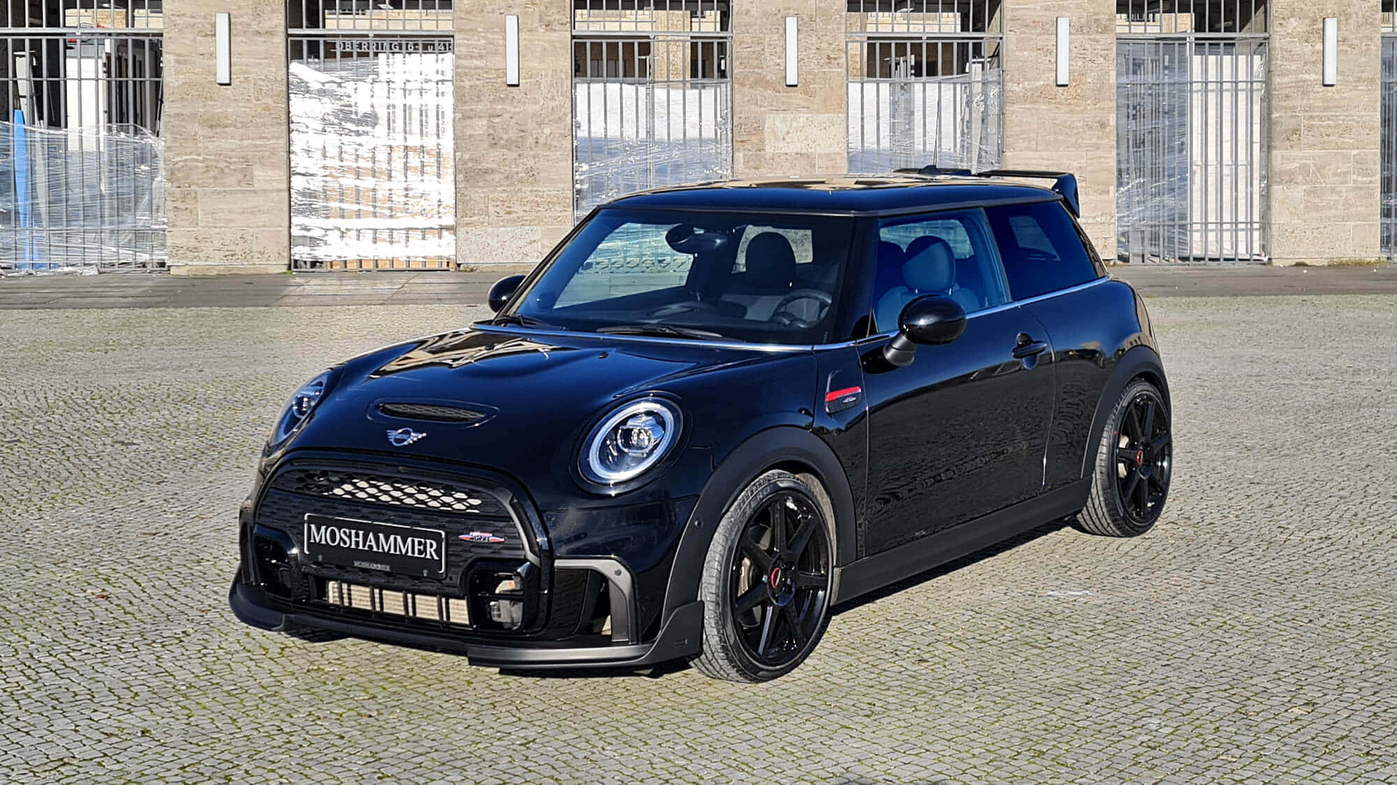 Mini Cooper S 桌布 壁紙 — 一輛亮黑色Moshammer改裝的Mini Cooper JCW停放在磚砌建築物前的鵝卵石庭院，建築物有金屬閘門。