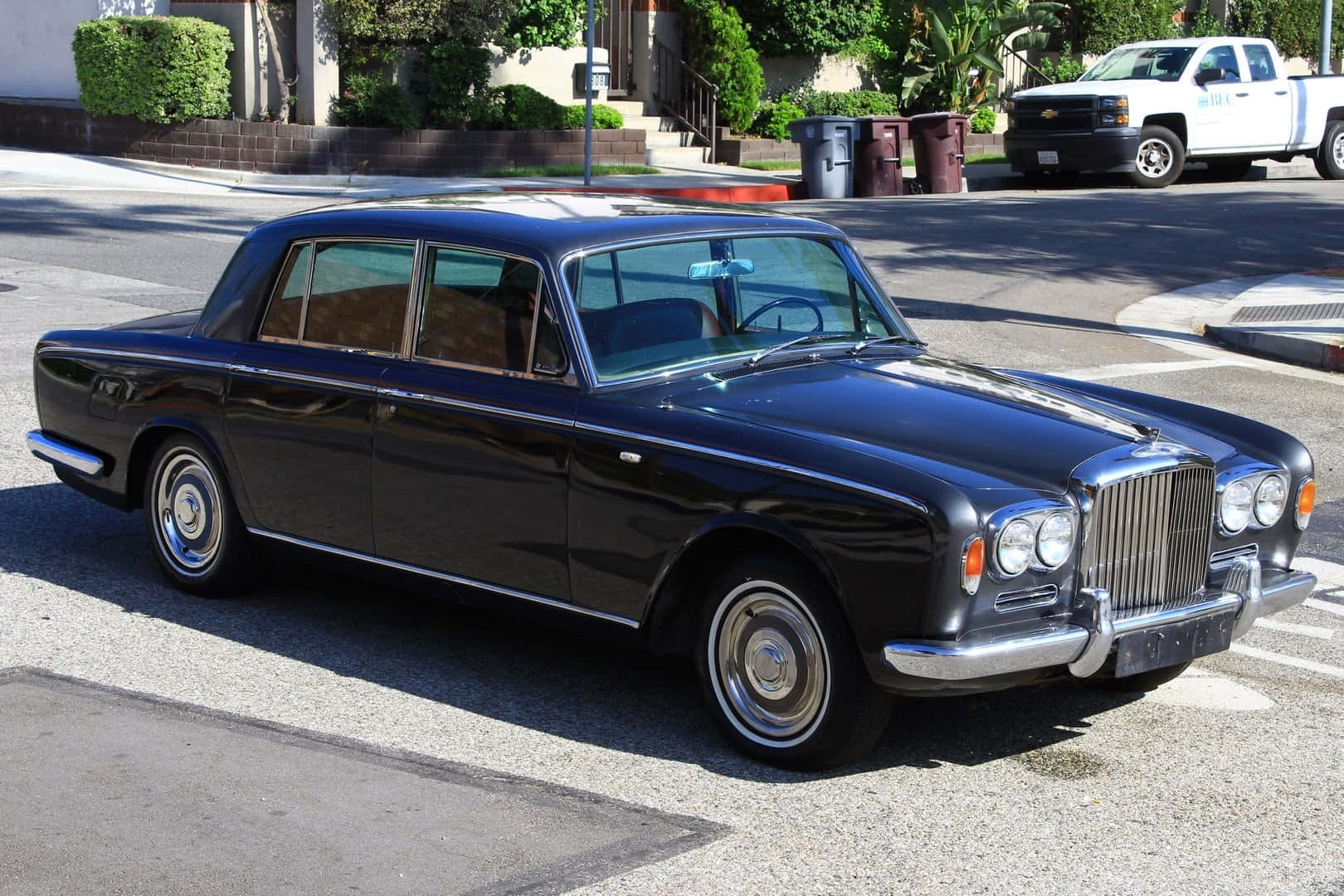 Bentley T1 桌布 壁紙 — 一輛亮黑色 vintage Bentley 房車停放在陽光普照的住宅街道上，背景有綠化和一輛白色卡車。