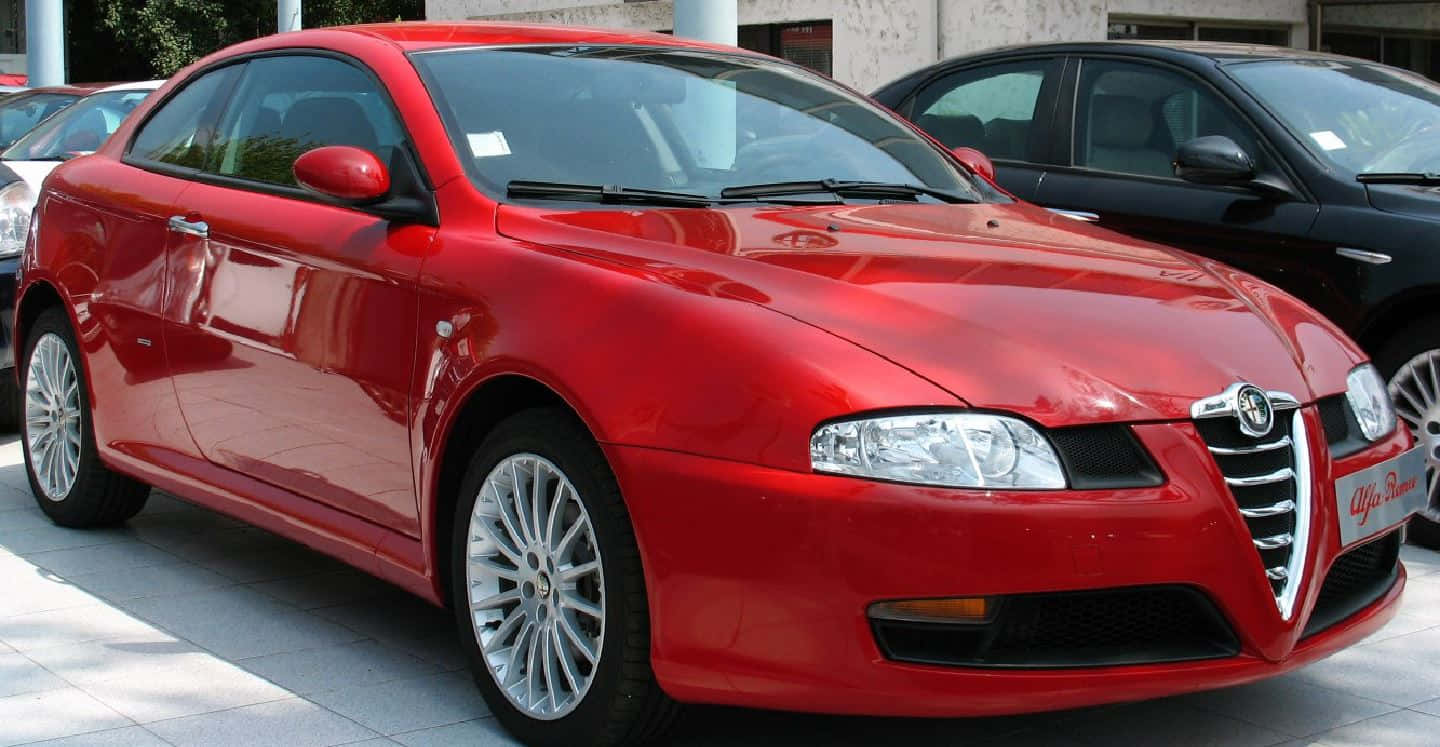 Alfa Romeo Gt 桌布 壁紙 — 一輛亮紅色的愛快羅密歐轎跑車在明亮的日光下閃閃發光，停泊在鋪有瓷磚的停車場上，附近有其他車輛。