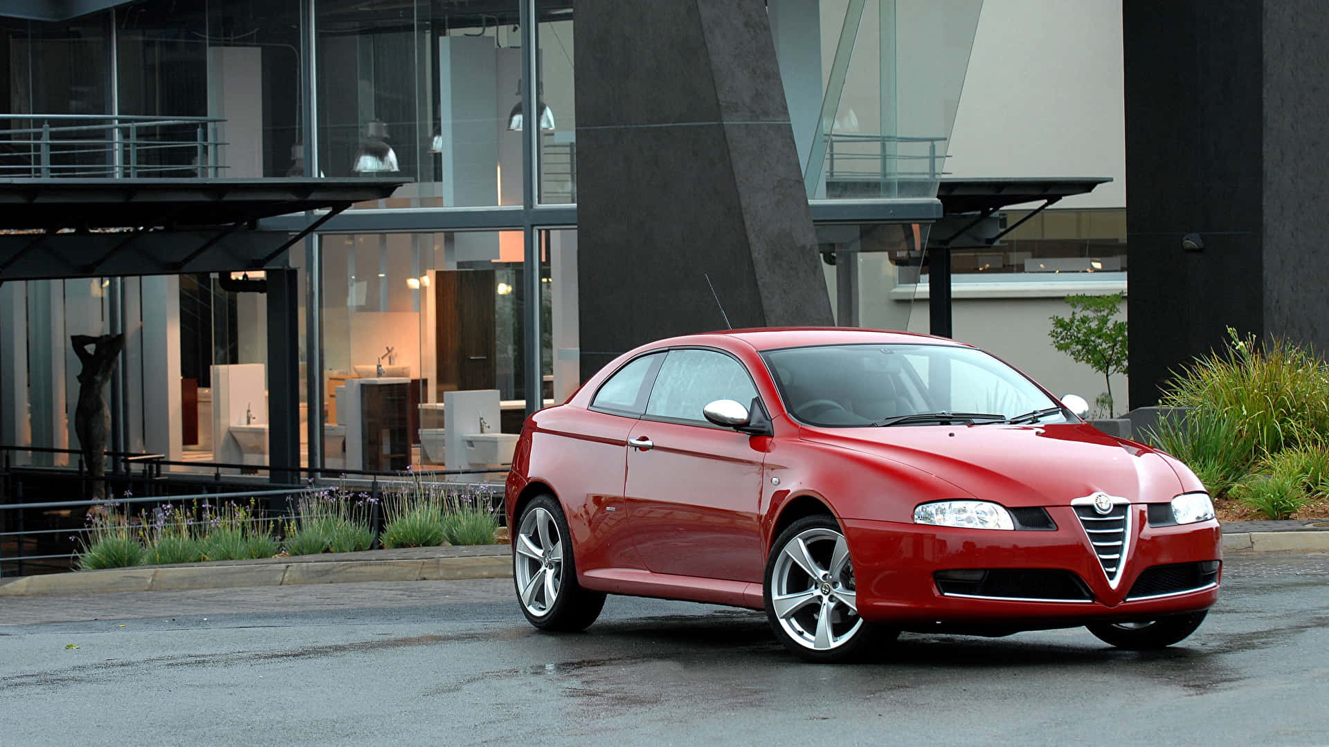 Alfa Romeo Gt 桌布 壁紙 — 一輛亮紅色的愛快羅密歐雙門跑車停泊在濕滑路面上，前方是一座現代玻璃建築，內部裝飾清晰可見。
