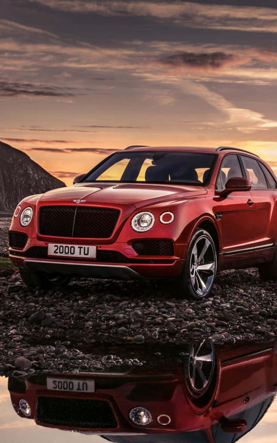 Bentley Bentayga 桌布 壁紙 — 一輛亮紅色的賓利Bentayga SUV停在鵝卵石上，背景是戲劇性的日落天空，下方水面映出倒影。