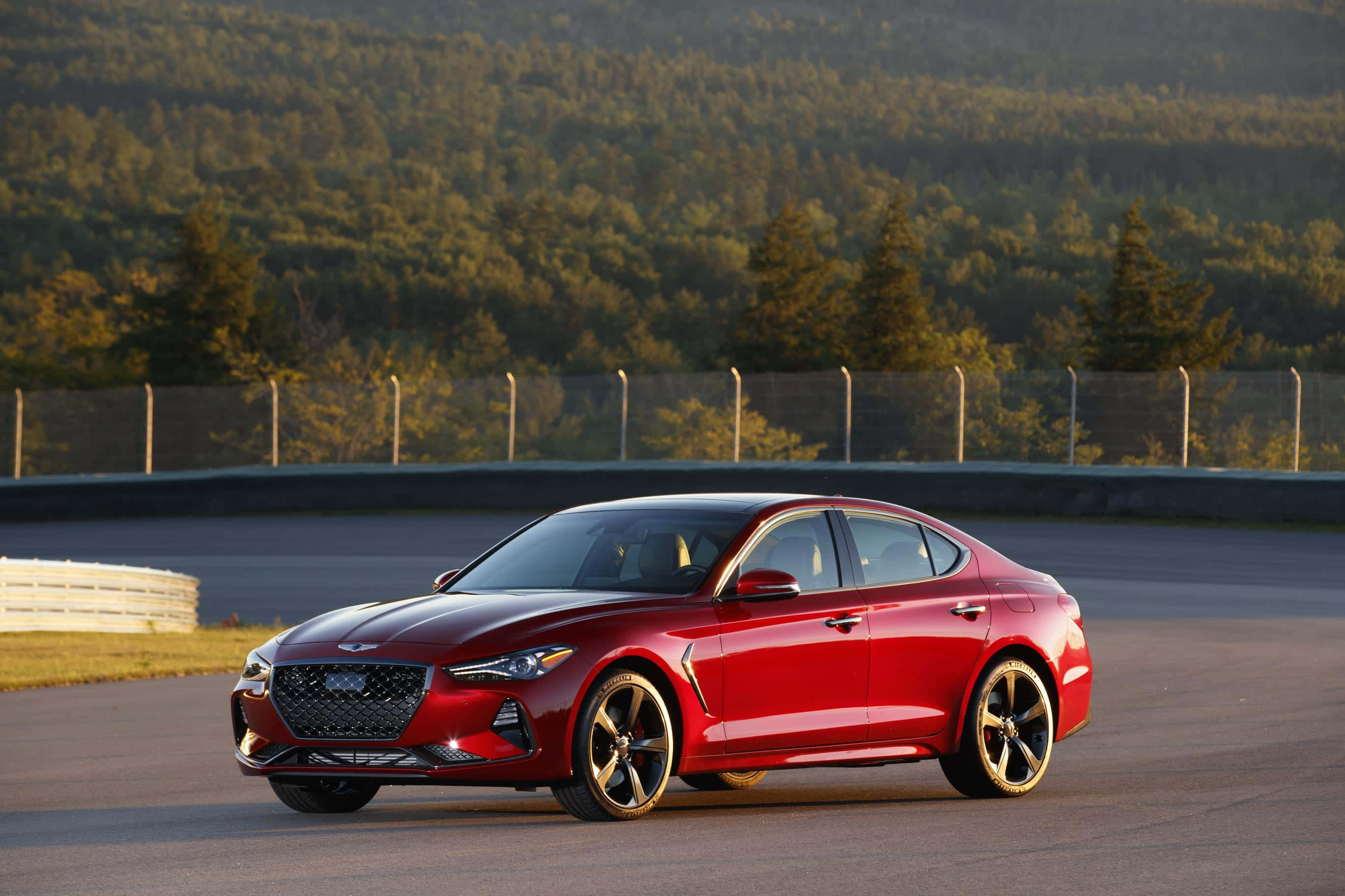 Genesis G70 桌布 壁紙 — 一輛亮紅色的跑車在黃金光線下，沿著被森林覆蓋的山丘環繞的賽道滑行。