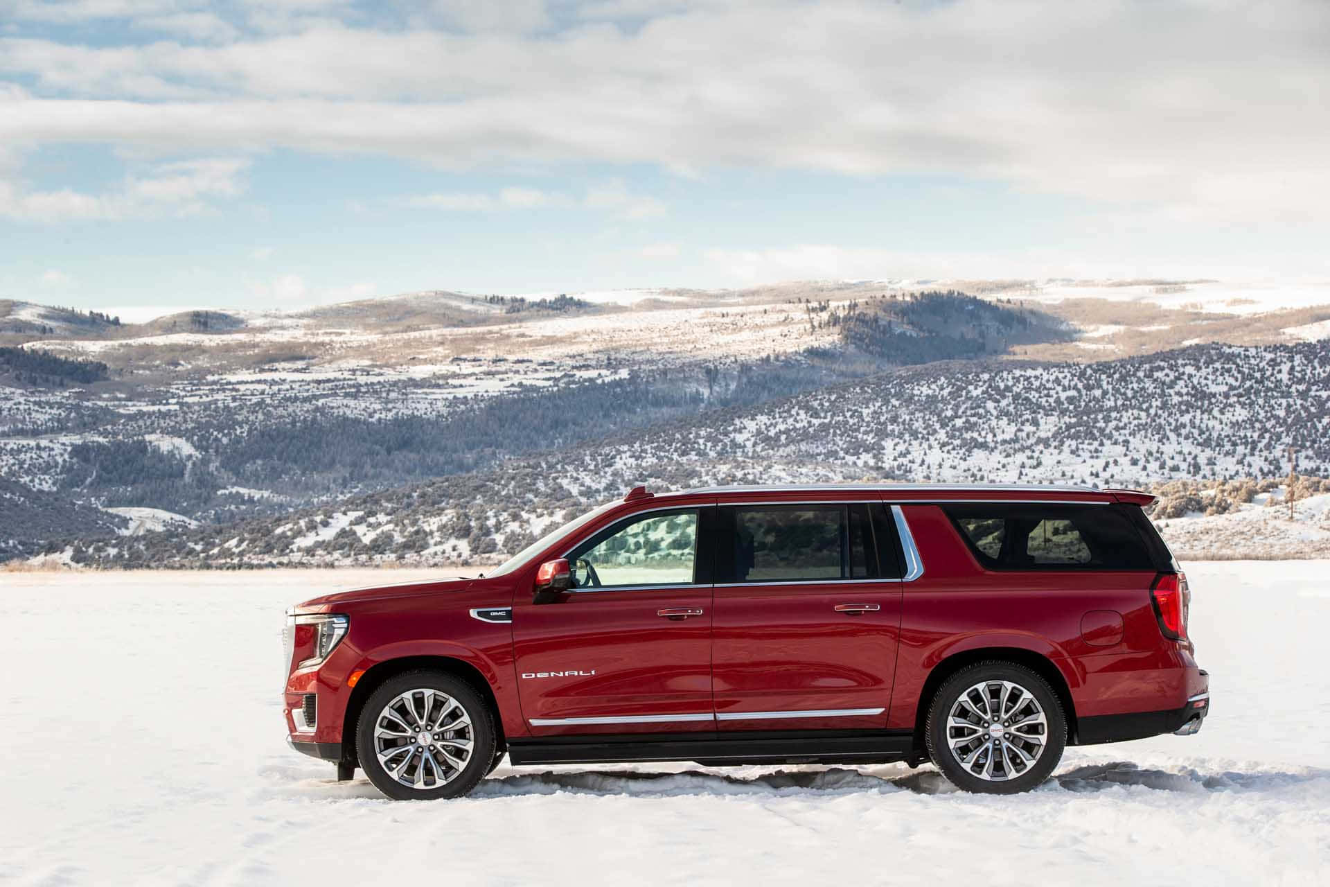 Gmc Yukon 桌布 壁紙 — 一輛亮紅色的Denali SUV停放在廣闊的雪地上，背景是部分多雲的天空下積雪的山巒。