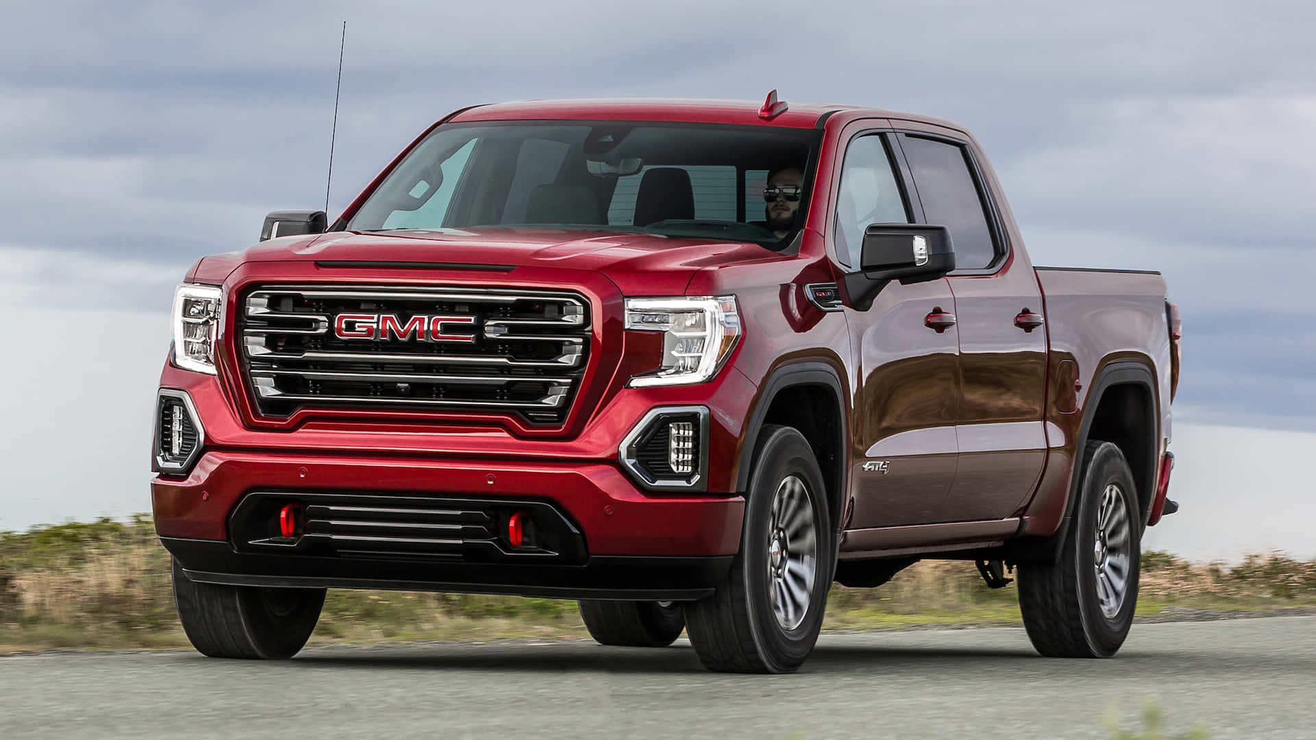 Gmc Sierra 桌布 壁紙 — 一輛亮紅色的GMC Sierra皮卡車在陰天下行駛在鋪砌的道路上，背景有乾草。