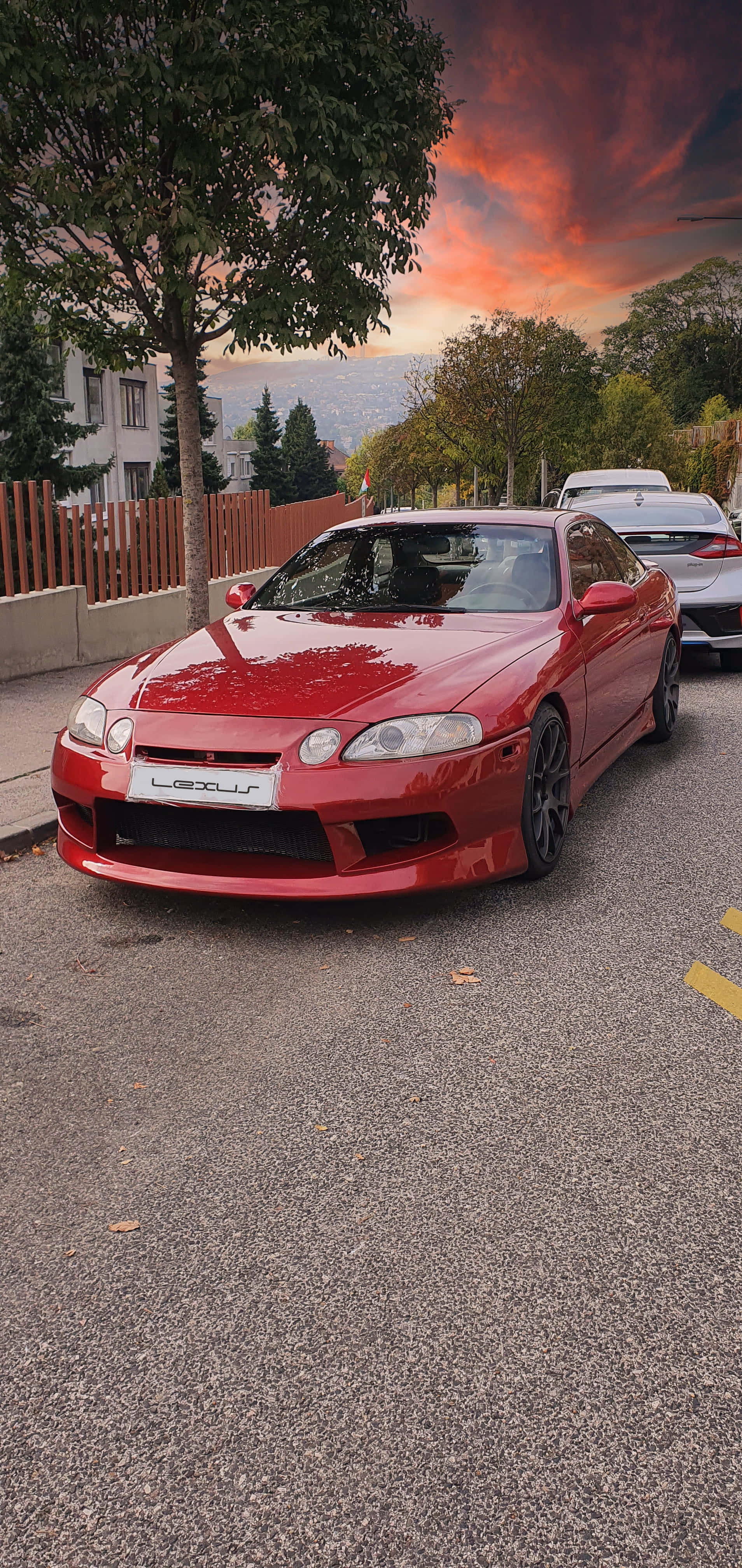 Lexus Sc 300 桌布 壁紙 — 一輛亮紅色Lexus跑車停泊在街上，背景是火燒般的夕陽天空，還有樹木和房屋。