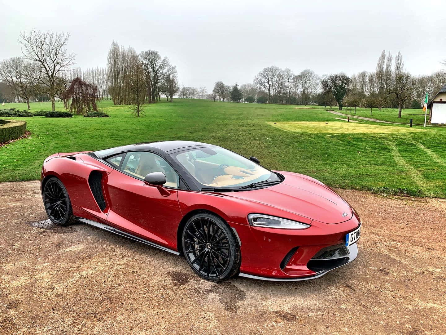 Mclaren Gt 桌布 壁紙 — 一輛亮紅色的McLaren跑車停在碎石車道上，旁邊是修剪整齊的綠色草坪和光禿禿的樹木，天空多雲。
