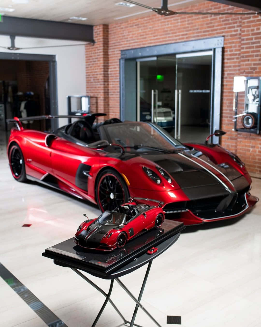 Pagani Huayra Roadster Bc 桌布 壁紙 — 一輛亮紅色的帕加尼風神跑車在現代車庫中閃耀，旁邊的展示架上放著同款比例模型。