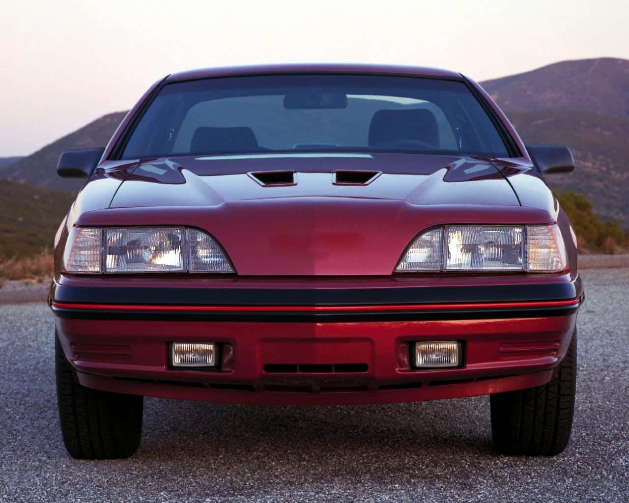 Ford Thunderbird 桌布 壁紙 — 一輛亮紅色跑車帶有引擎蓋進氣口，在碎石路上向前行駛，背景是日落時分的連綿山丘。