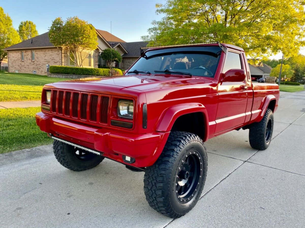 Jeep Comanche 桌布 壁紙 — 一輛亮紅色升高底盤的Jeep Cherokee皮卡車停在混凝土車道上，背景是郊區房屋和綠樹，天空清澈湛藍。