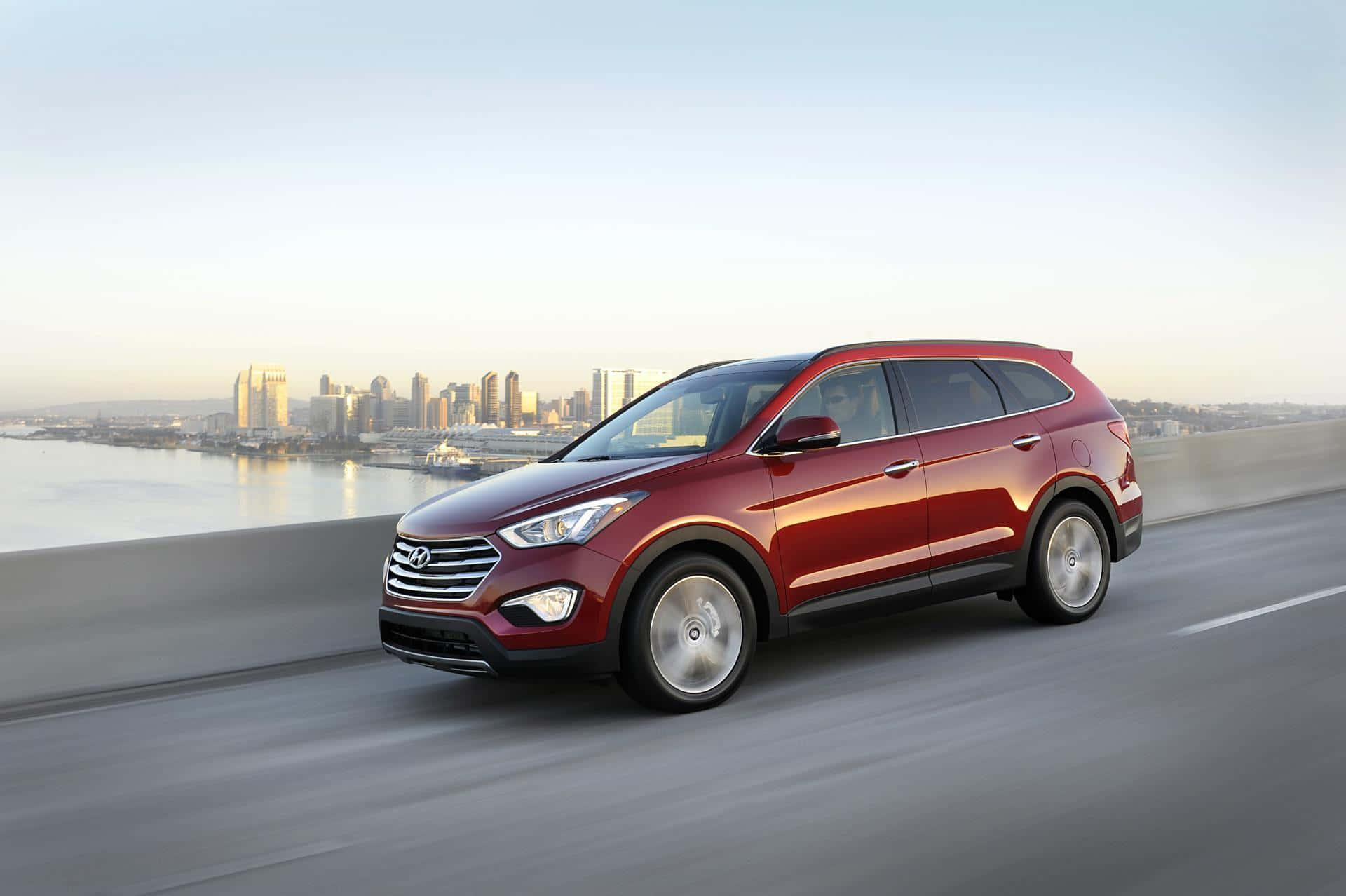 Hyundai Santa Fe 桌布 壁紙 — 一輛亮紅色SUV在高速公路上行駛，背景是黃金時段的城市天際線和海濱。
