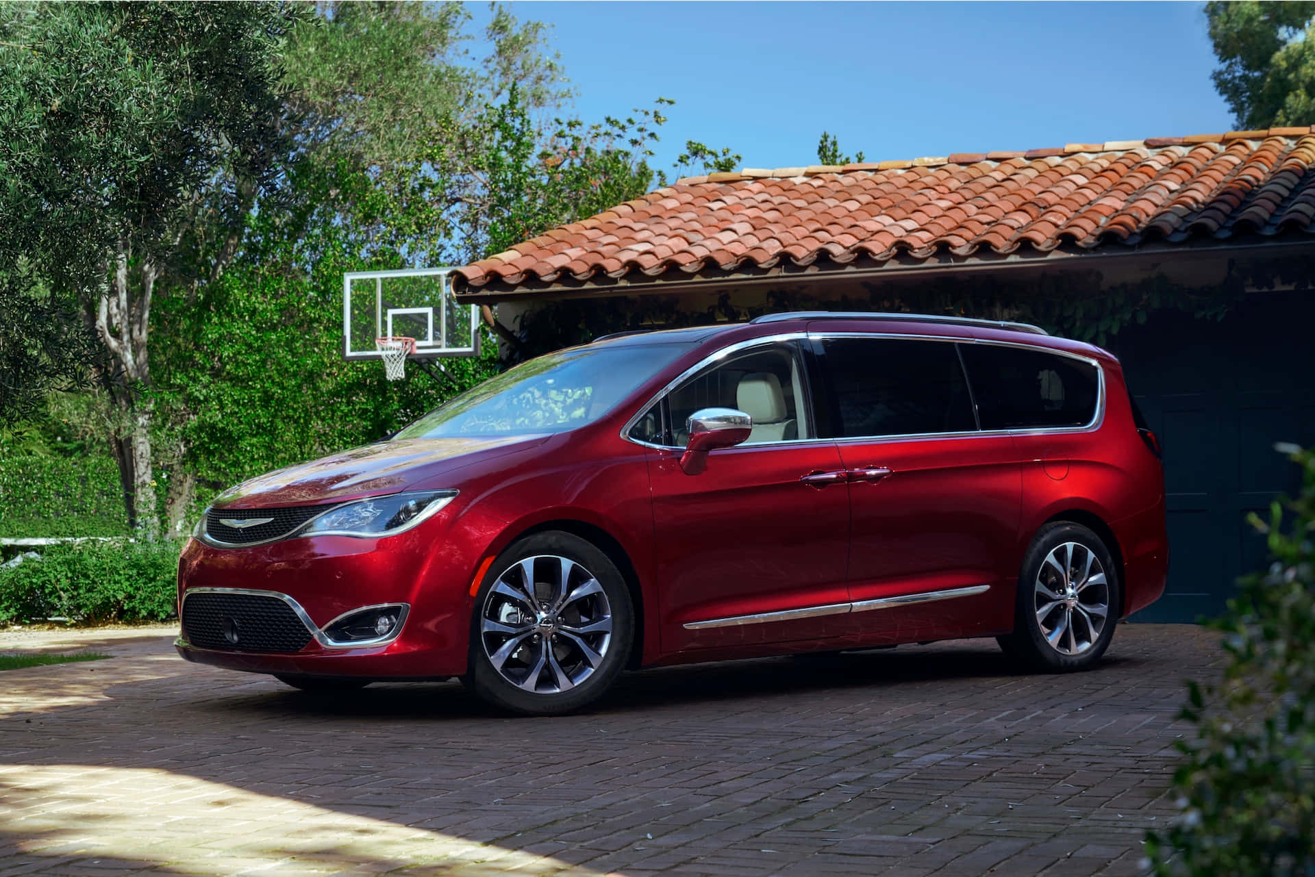 Chrysler Pacifica 桌布 壁紙 — 一輛亮紅色小型貨車停在磚砌車道上，旁邊是瓷磚屋頂車庫，車庫上有籃球架，周圍是茂密的綠色庭院。