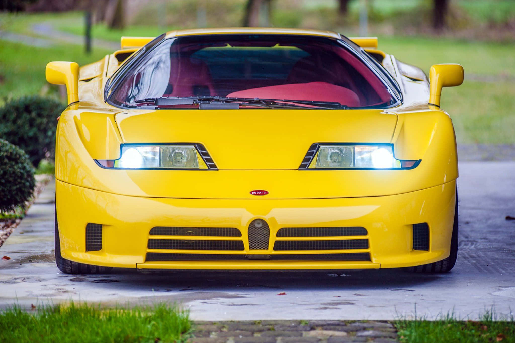 Bugatti Eb110 桌布 壁紙 — 一輛亮黃色的Bugatti EB110超級跑車，亮著車頭燈，停放在濕漉漉的車道上，周圍環繞著綠草和樹木。