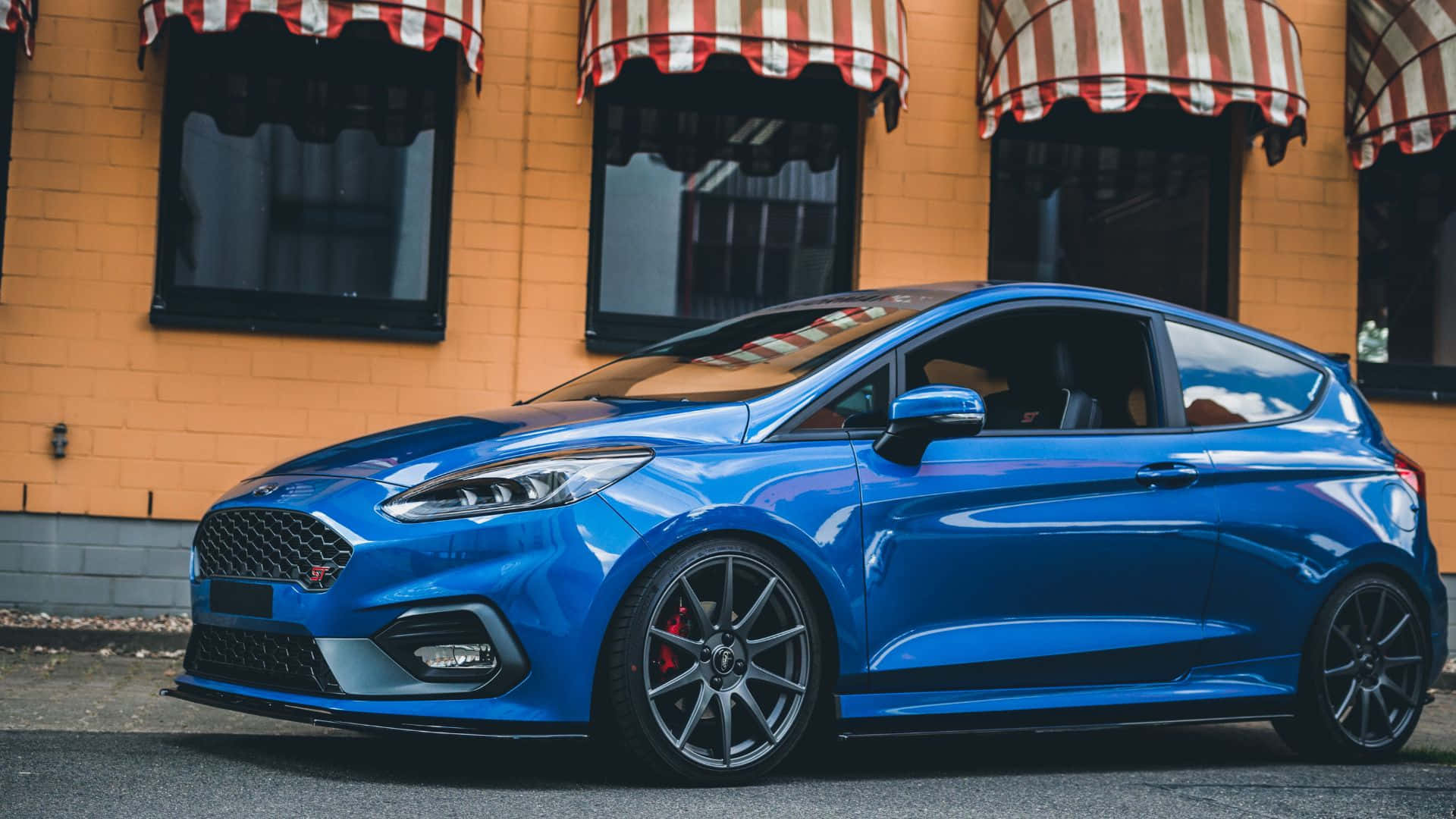 Ford Fiesta 桌布 壁紙 — 一輛亮藍色的福特 Fiesta ST 停在一棟黃色磚砌建築前，建築物有紅白條紋的遮陽篷。