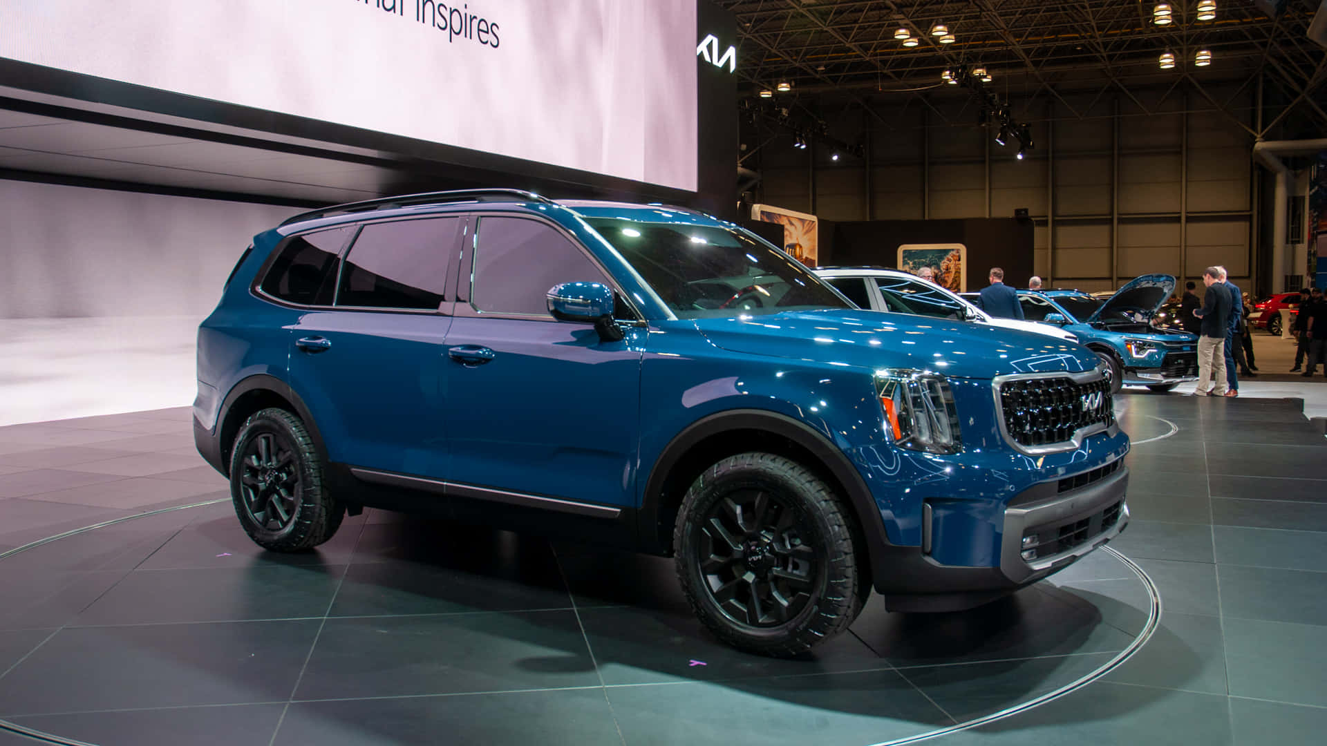 Kia Telluride 桌布 壁紙 — 一輛亮藍色Kia SUV在圓形平台上於車展中展示，背景有明亮的頭頂燈光和與會者。