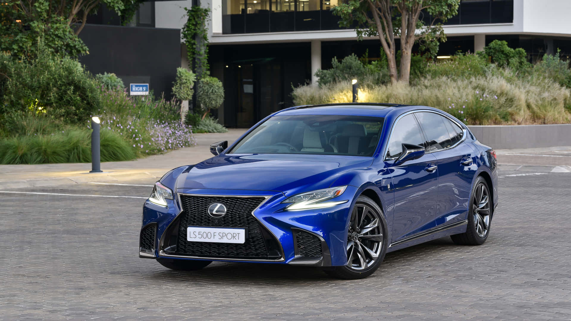 Lexus Ls 500 桌布 壁紙 — 一輛亮藍色Lexus LS 500 F SPORT轎車停放在鋪砌的車道上，旁邊是現代建築和綠植。