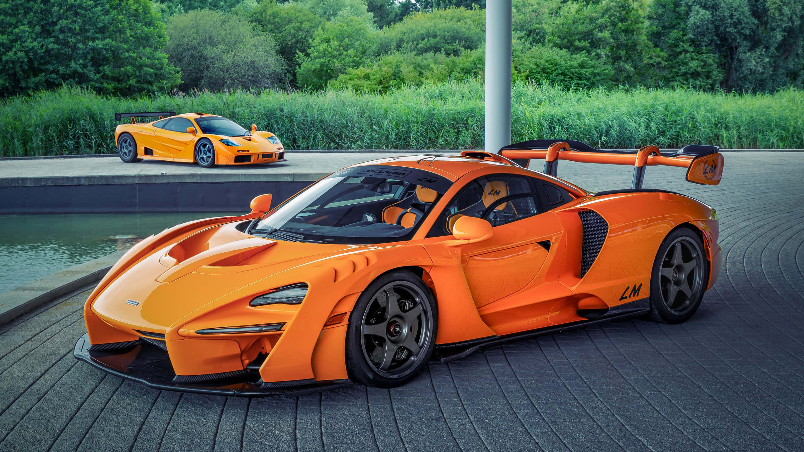 Mclaren F1 桌布 壁紙 — 兩輛充滿活力的橙色McLaren超級跑車在日光下閃閃發光，旁邊是時尚的水景，周圍環繞著茂密的綠植。