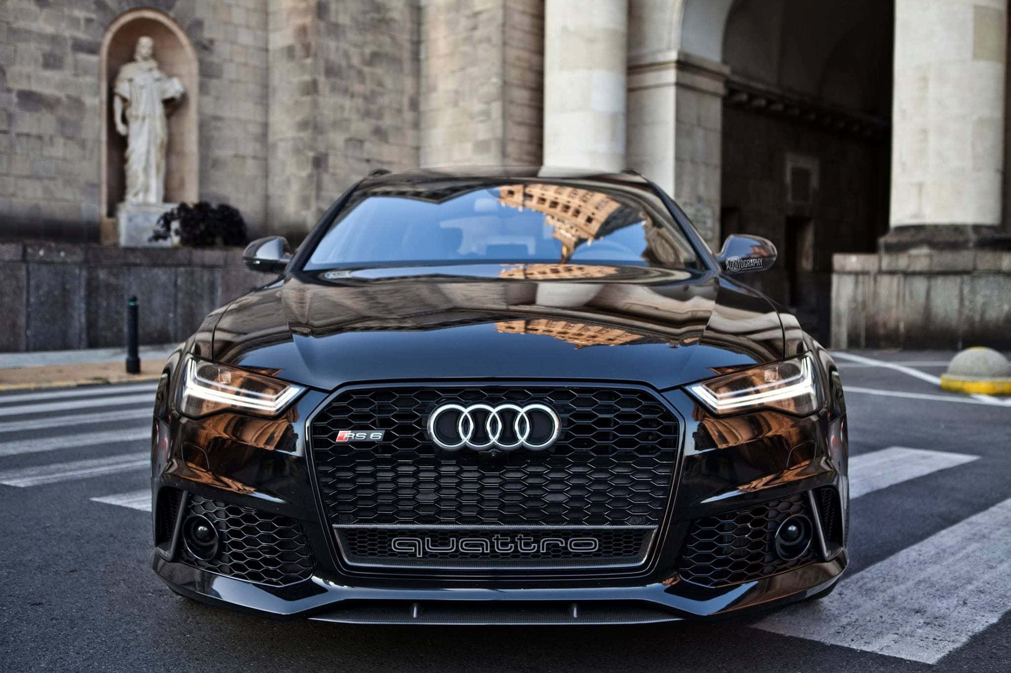 Audi Rs6 桌布 壁紙 — 一輛亮黑色的奧迪RS7跑車停泊在歷史石建築和雕像背景的城市街道上。