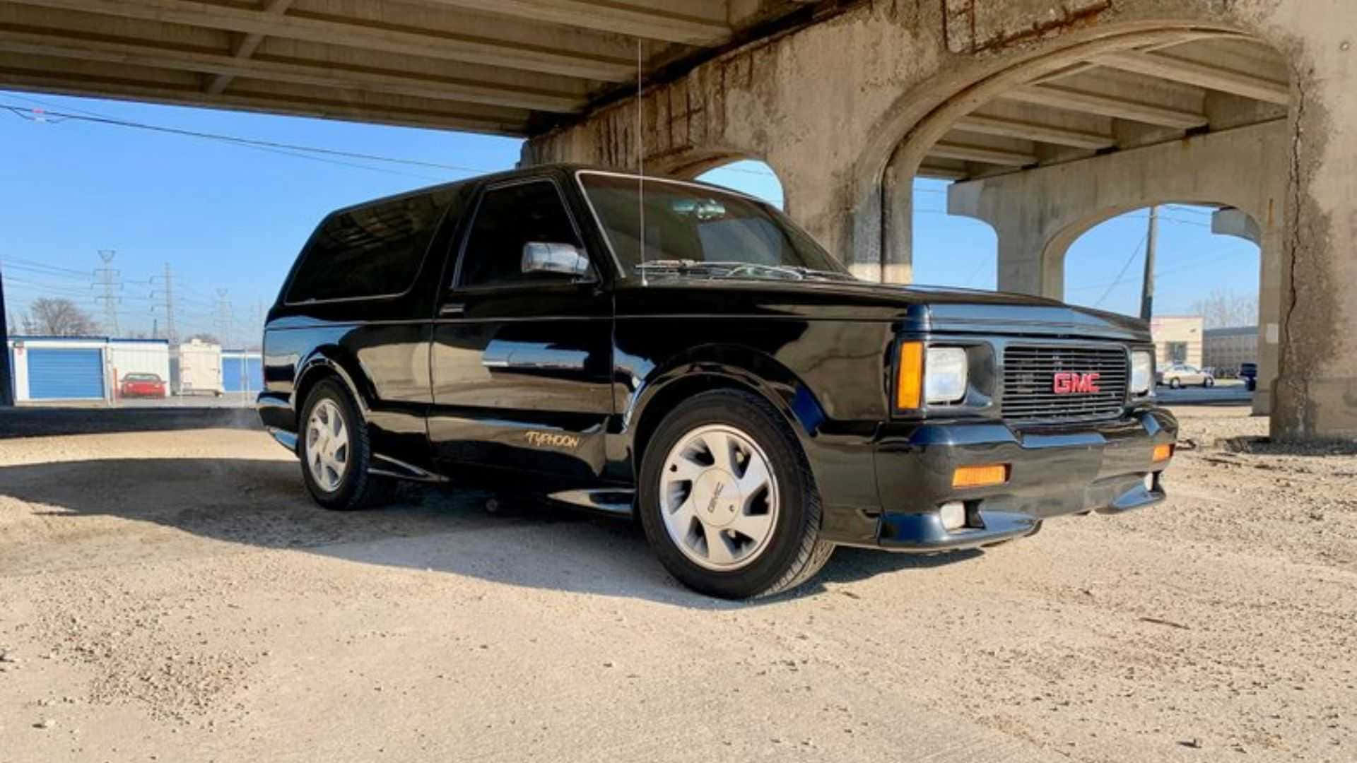 Gmc Typhoon 桌布 壁紙 — 一輛亮黑色GMC Typhoon SUV在晴朗天氣下停泊在風化混凝土天橋下。