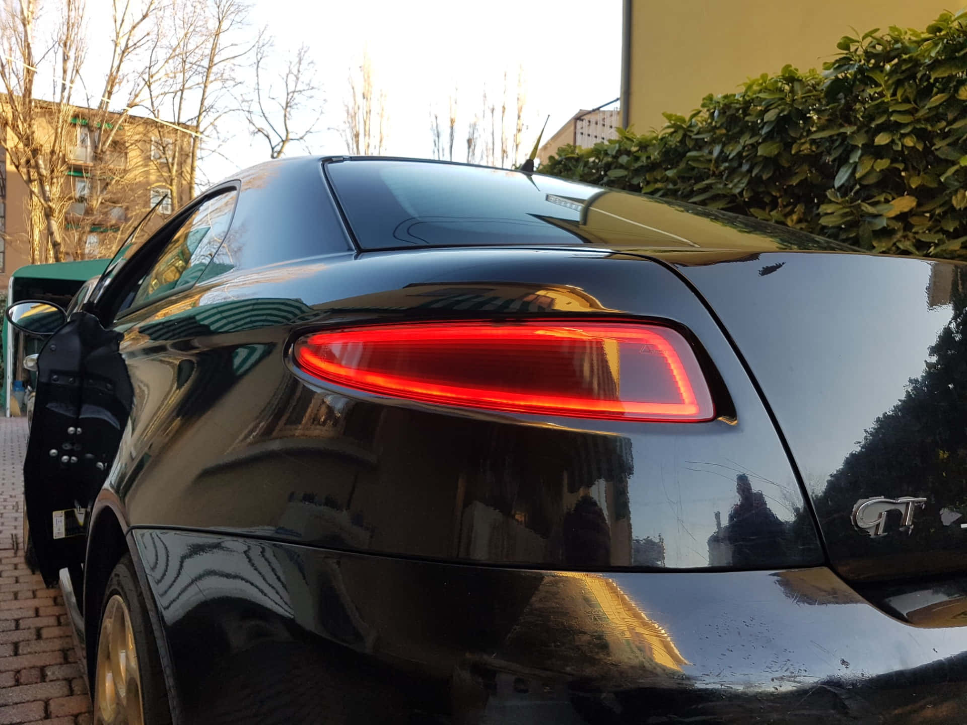 Alfa Romeo Gt 桌布 壁紙 — 一輛亮黑色跑車，帶有發光的紅色尾燈，停在磚街上，旁邊有綠色灌木和光禿禿的樹木。