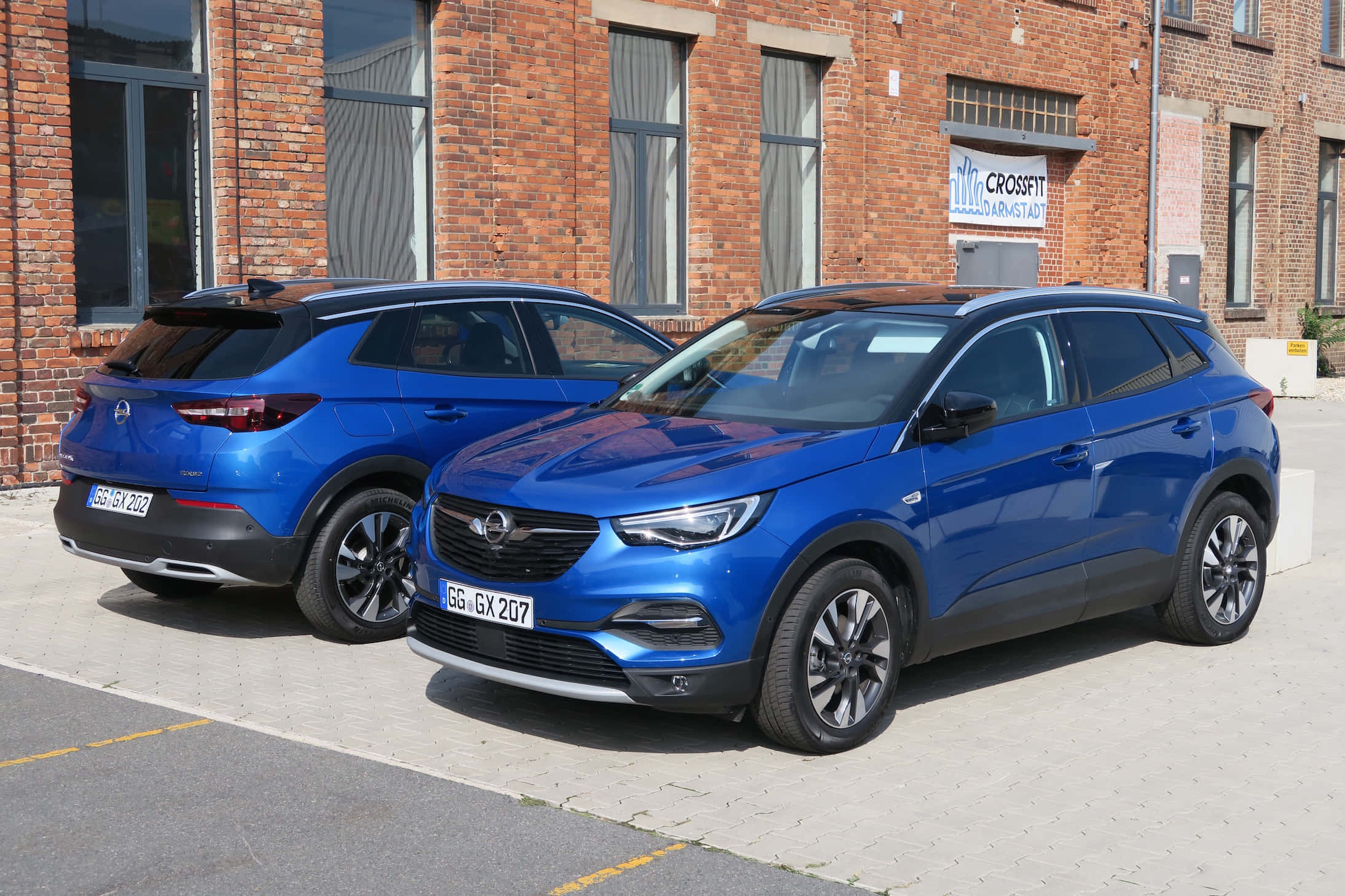 Opel Grandland X 桌布 壁紙 — 兩輛亮藍色歐寶SUV並排停放在紅磚建築前的鋪砌場地上。