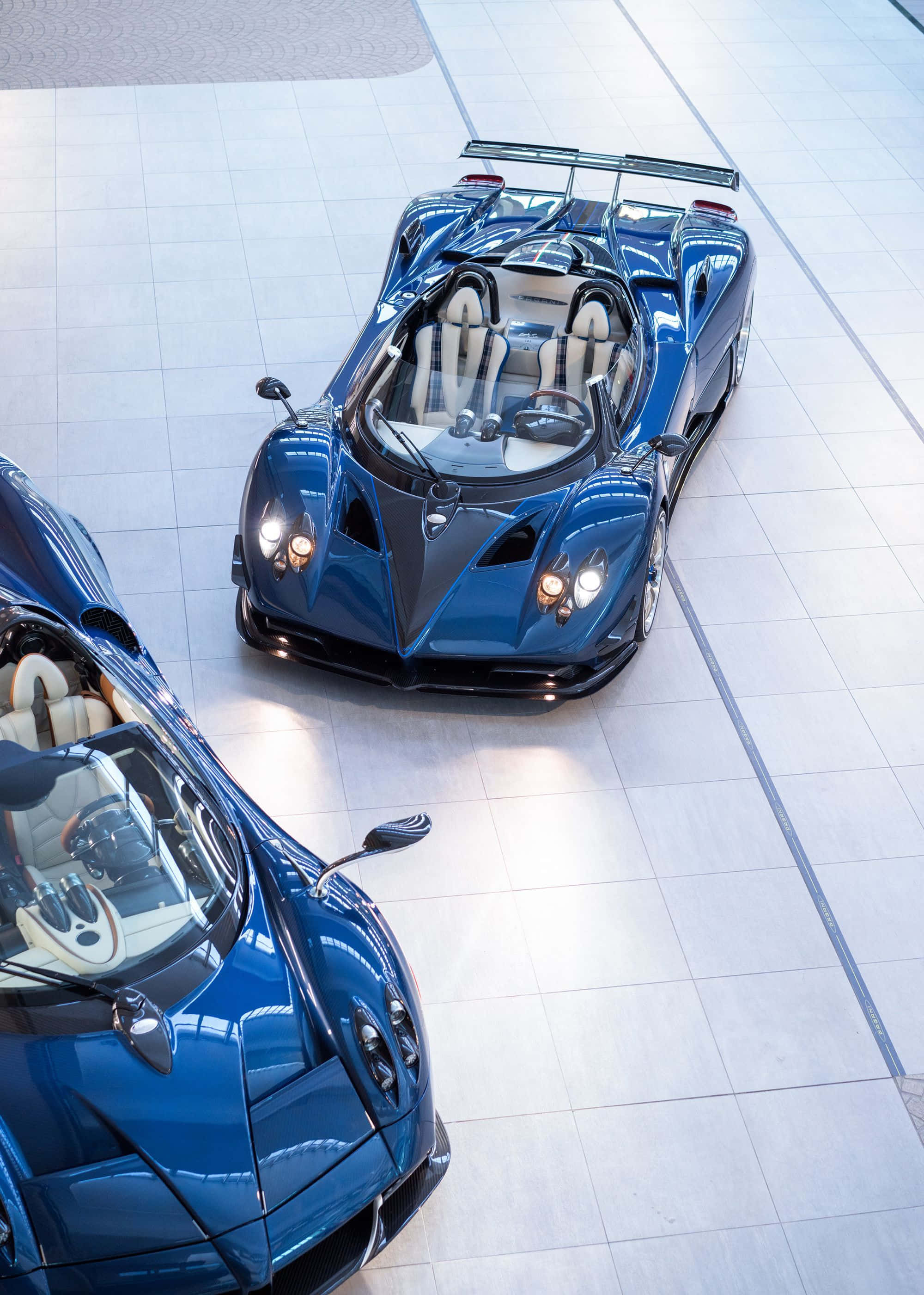 Pagani Zonda Hp Barchetta 桌布 壁紙 — 兩輛線條流暢的藍色超級跑車，白色內裝，在拋光瓷磚地板上閃爍著明亮燈光。