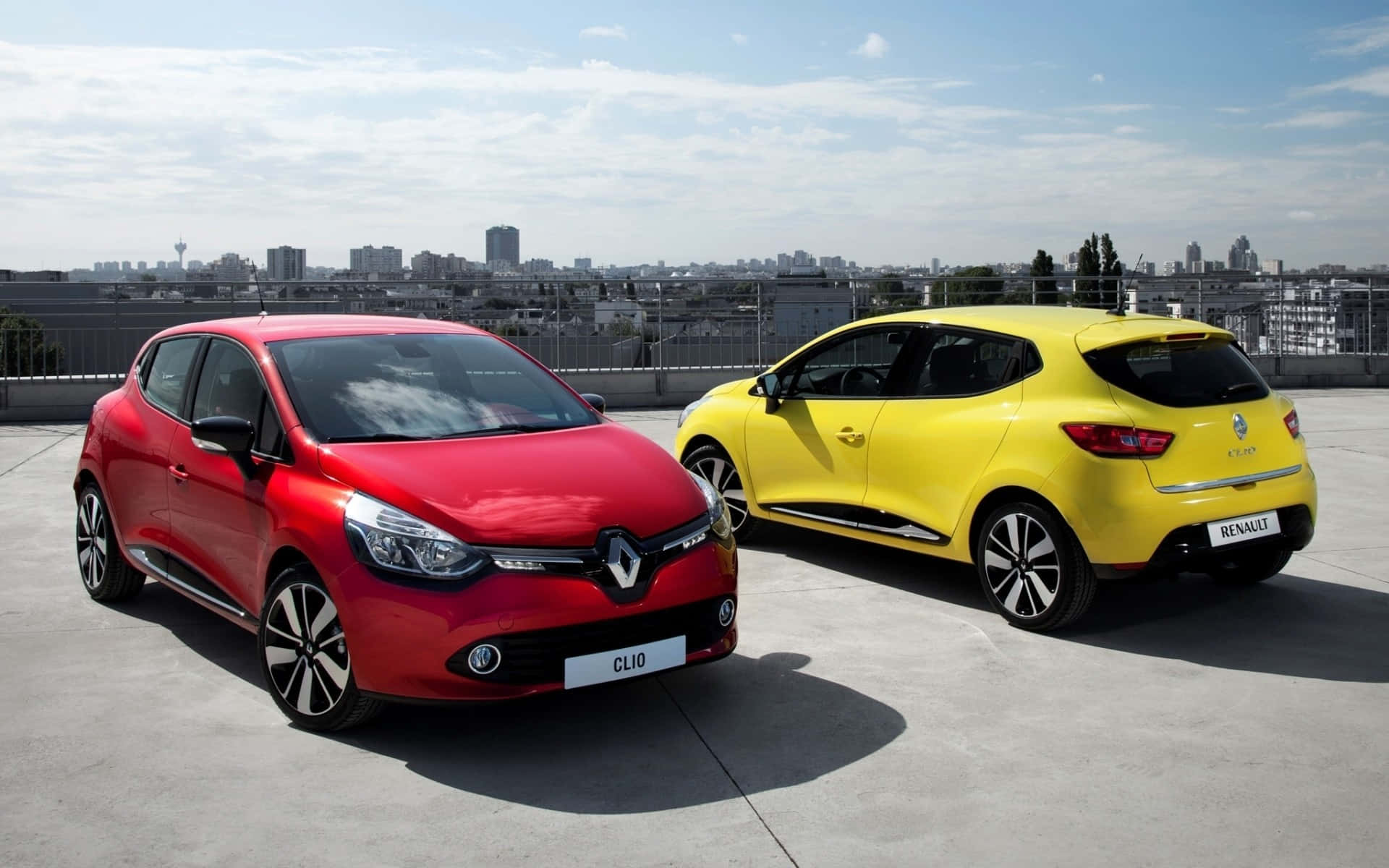 Renault Clio 桌布 壁紙 — 兩輛鮮豔的雷諾汽車紅色和黃色停放在屋頂上，背景是城市天際線，天空湛藍。