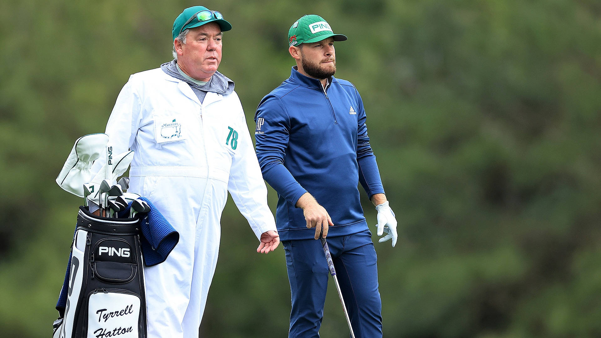 Tyrrell Hatton 桌布 壁紙 — 兩名戴綠色帽的高爾夫球手並肩走在翠綠的球場上，其中一人背著印有Tyrrell Hatton標籤的PING球袋。