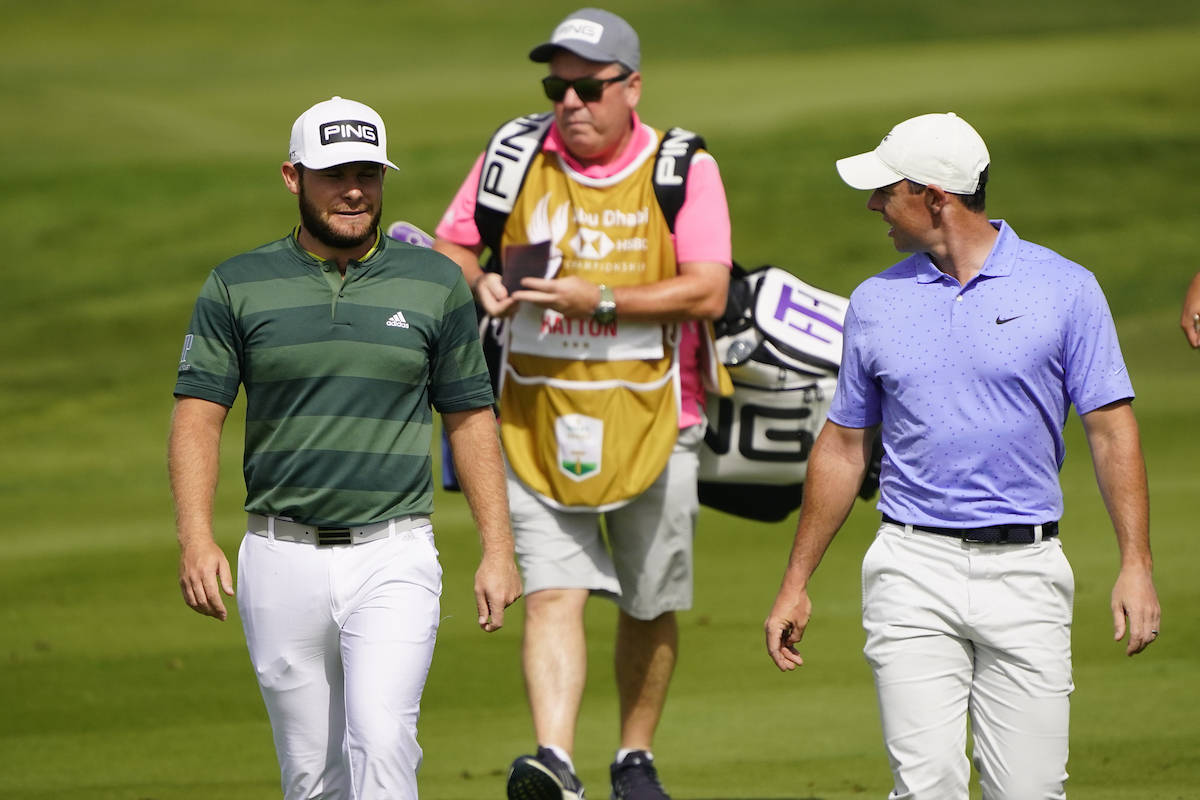 Tyrrell Hatton 桌布 壁紙 — 兩名高爾夫球手穿著整潔的服裝，在陽光照耀的綠色球道上行走，身後跟著一名球僮。