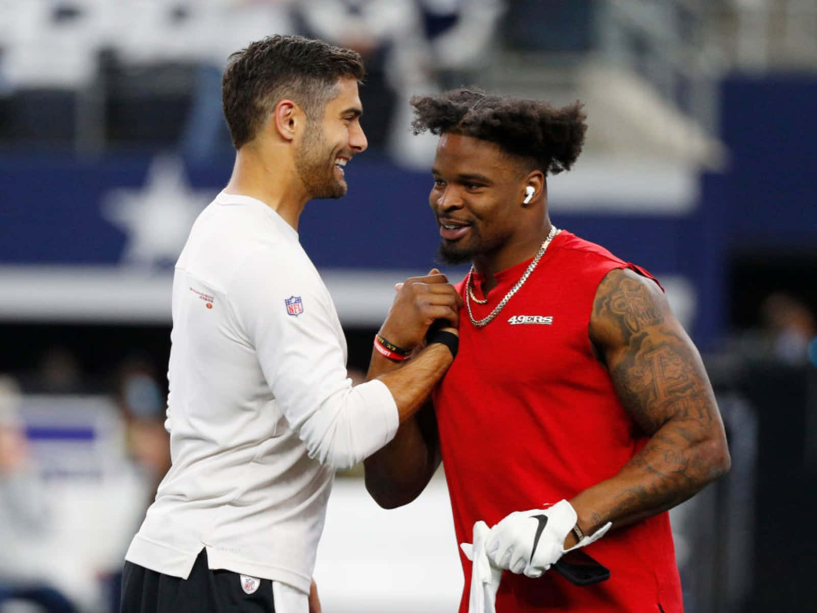  壁紙 — 兩名面帶微笑的NFL球員身穿隊服，在明亮的燈光下於體育場上友好地碰拳。