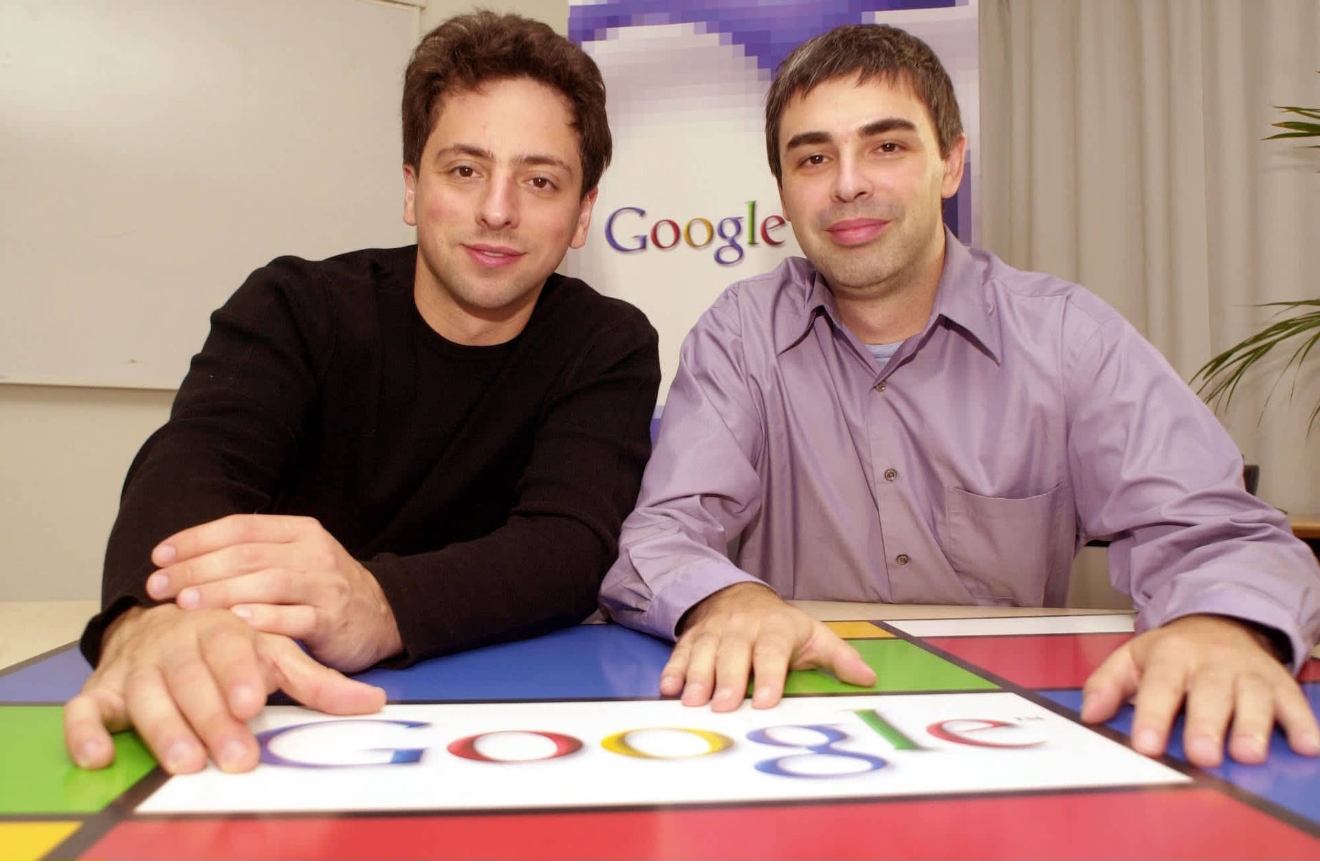 兩名男士在辦公室Google標誌辦公桌後對著鏡頭微笑 — Google 壁紙