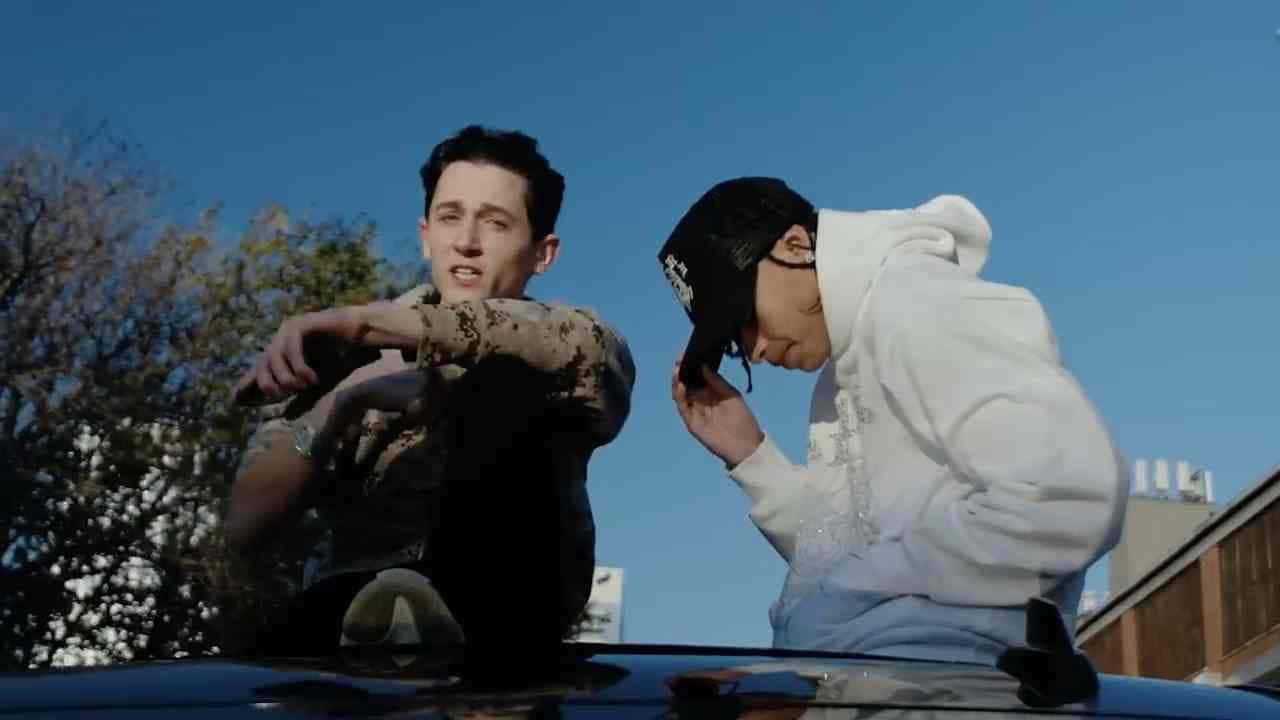  壁紙 — 兩名年輕男子在晴朗藍天下靠著車頂，背景有樹木和建築物。