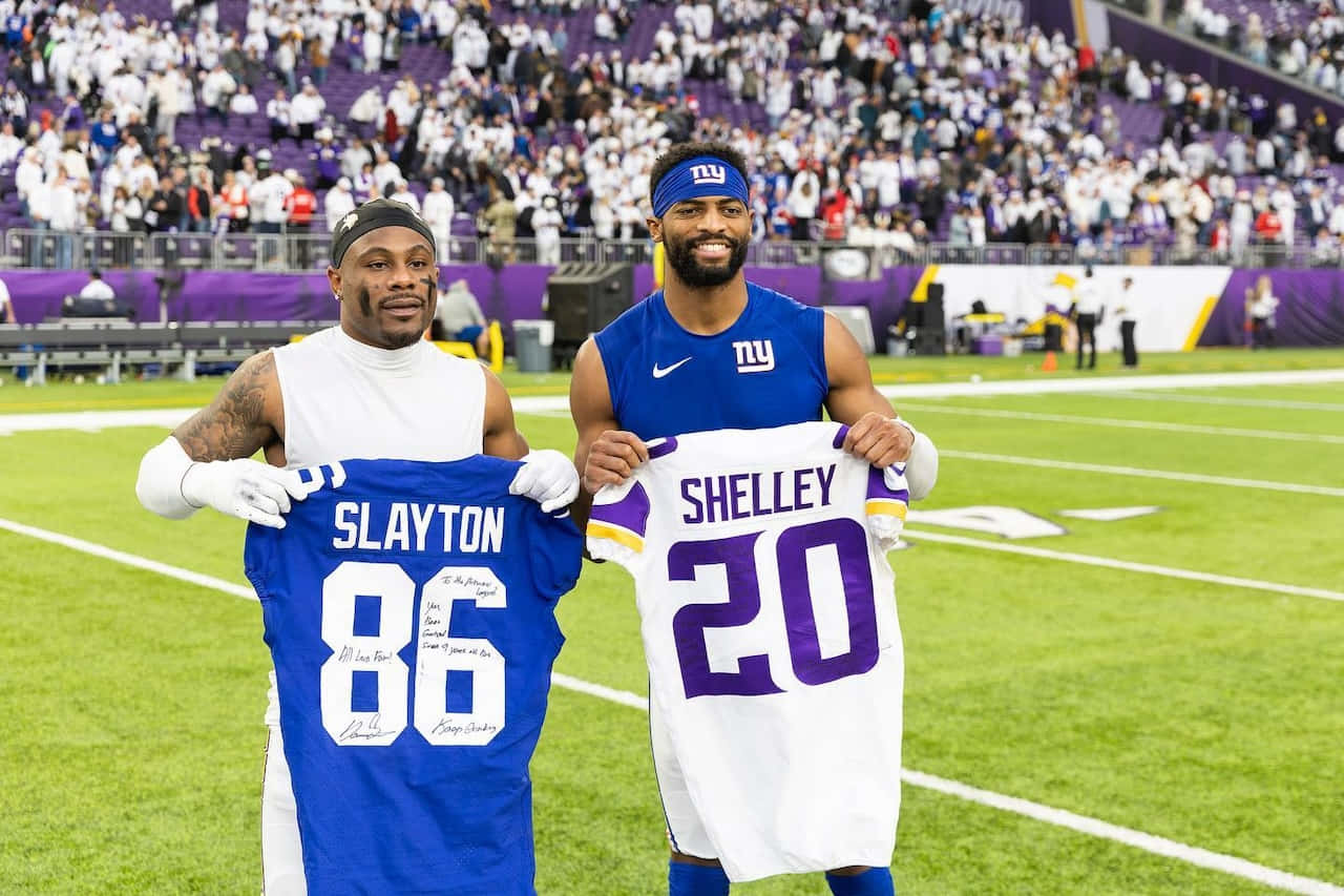 壁紙 — 兩名微笑的NFL球員在綠色球場上，在座無虛席的體育館前舉起帶有名字和號碼的簽名球衣。