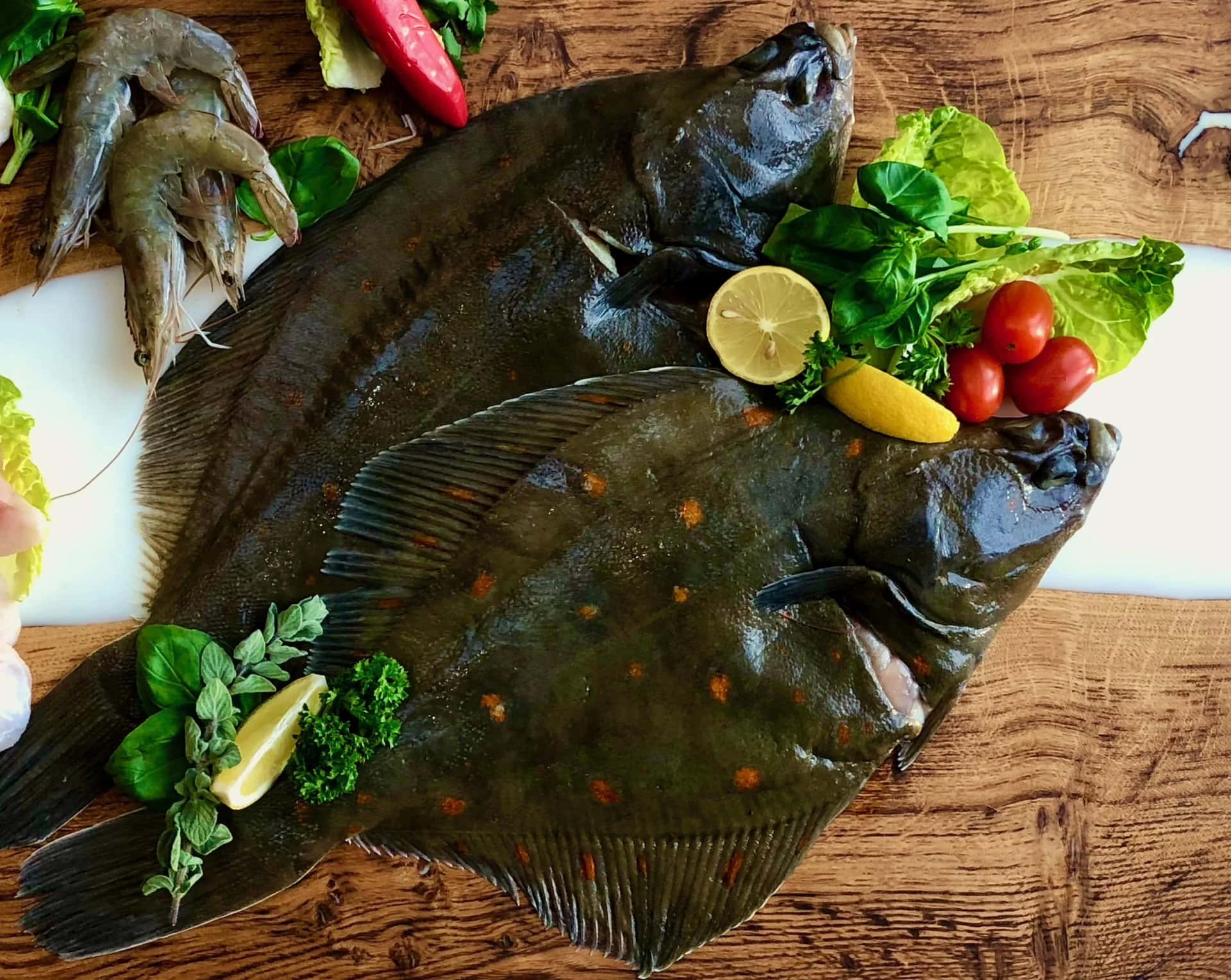  壁紙 — 兩條新鮮的比目魚放在木板上，周圍有蝦、小番茄、檸檬片和新鮮香草。