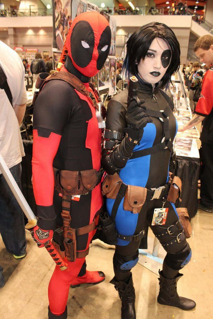 Deadpool And Domino 桌布 壁紙 — 兩位穿著精緻死侍和黑寡婦服裝的角色扮演者在熱鬧的漫畫展上一起擺姿勢。