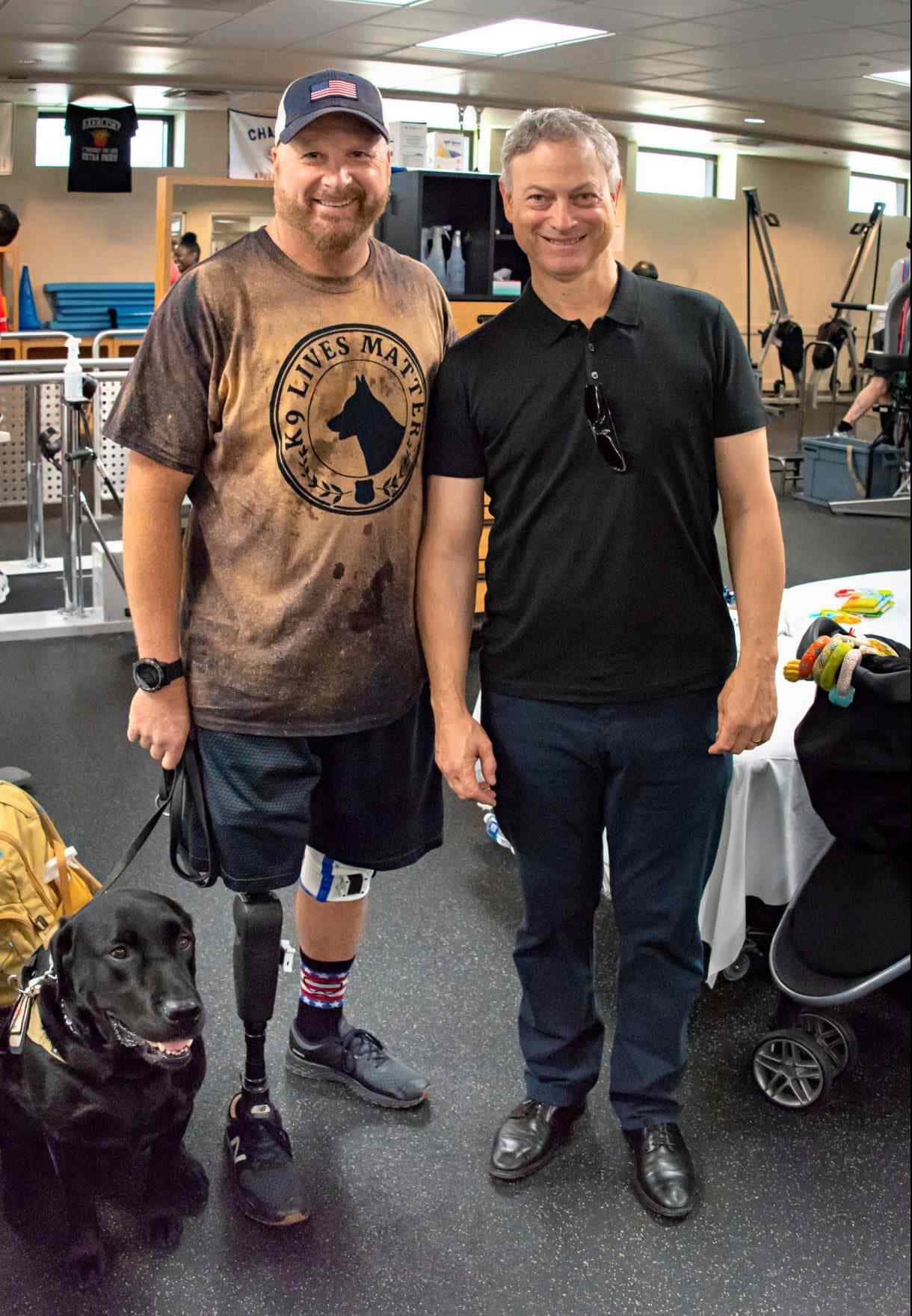  壁紙 — 兩位微笑的男子在類似健身房的環境中，與一隻黑色服務犬和附近的嬰兒車一起站在室內。