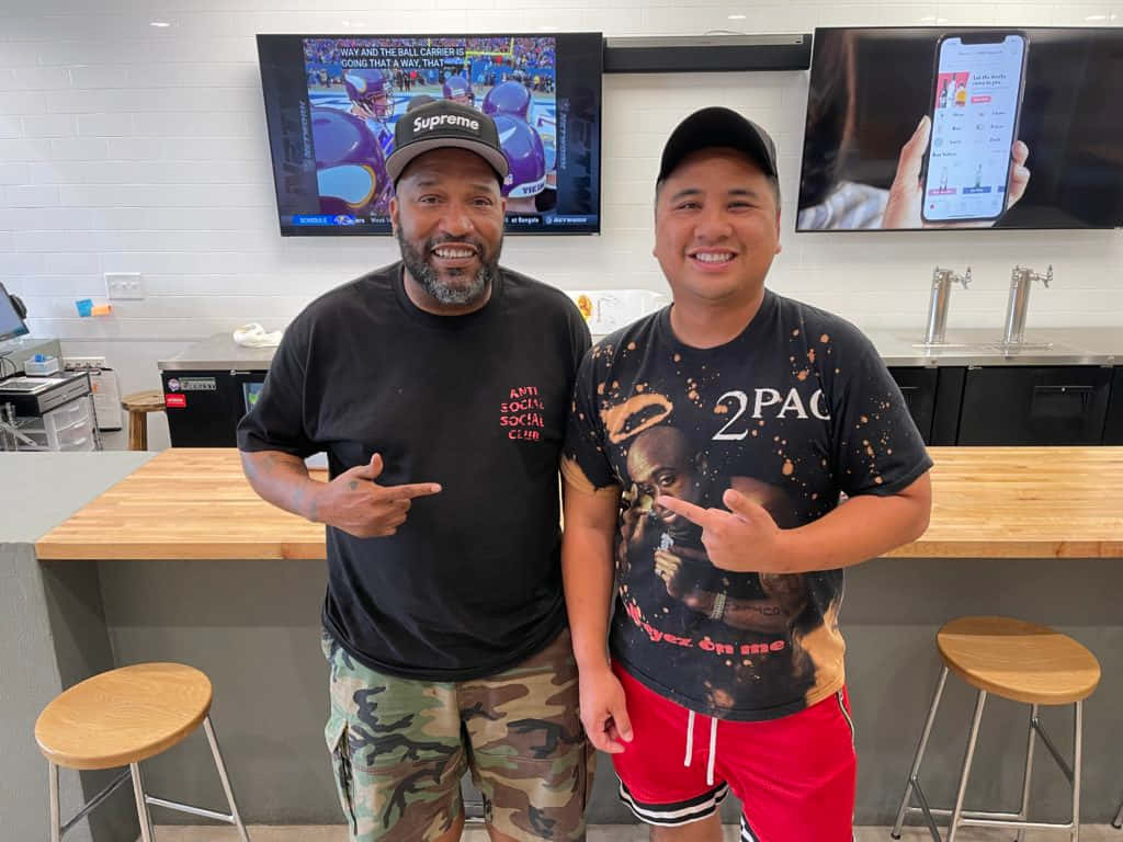  壁紙 — 兩位微笑的男子在現代酒吧中擺姿勢，身後有顯示體育賽事的電視螢幕和手機應用程式。