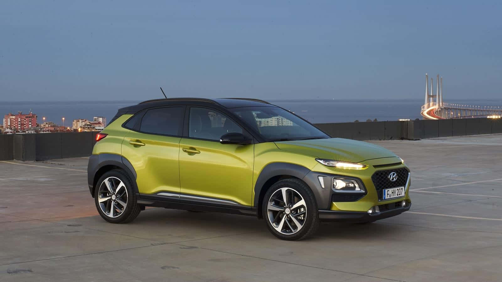 Hyundai Kona 桌布 壁紙 — 一輛鮮豔的青檸綠色現代Kona SUV停在海邊的觀景點，遠處有一座亮燈的橋樑。