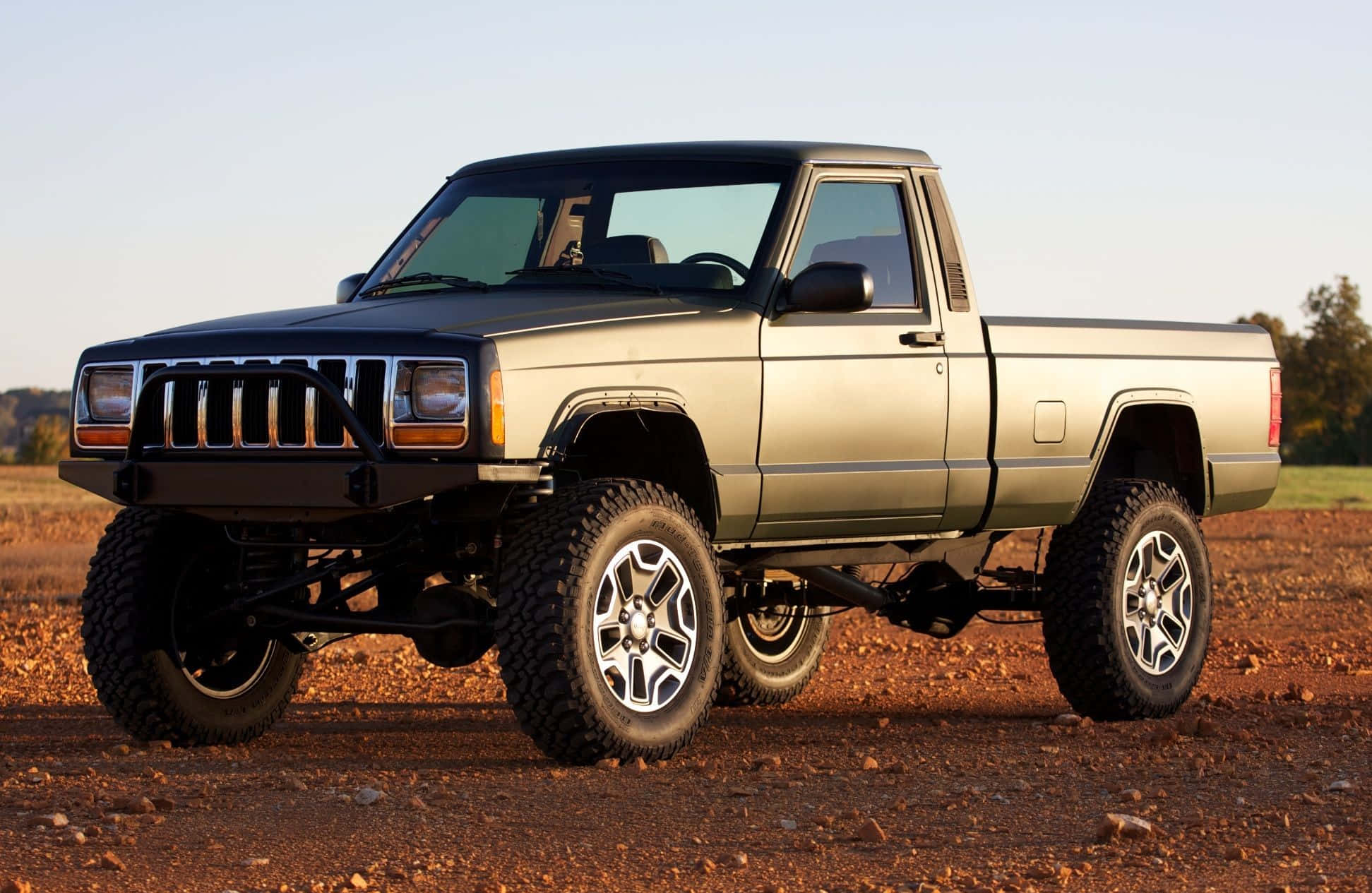 Jeep Comanche 桌布 壁紙 — 一輛升高了的銀色Jeep Cherokee皮卡車，配備大輪胎，在晴朗的天空下停在一片泥地上。