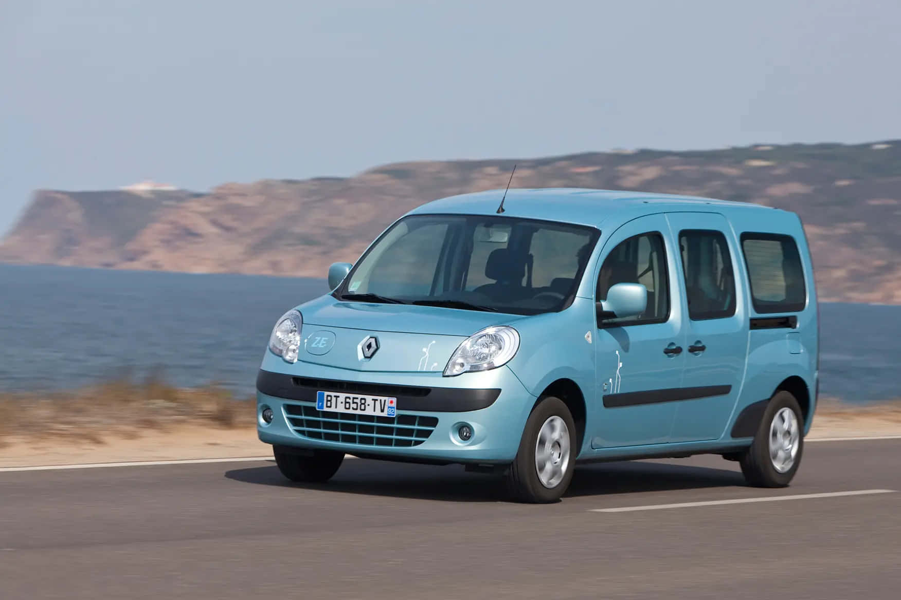 Renault Kangoo 桌布 壁紙 — 一輛淺藍色的雷諾 Kangoo 廂型車沿著海岸道路行駛，背景有懸崖和大海。