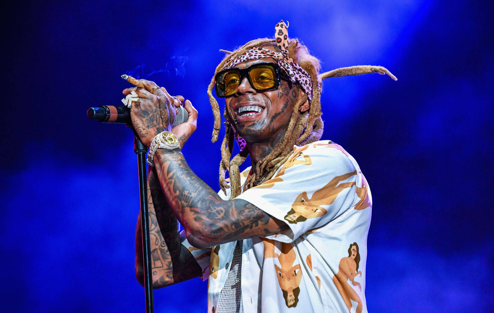 Lil Wayne 桌布 壁紙 — Lil Wayne 在舞台上穿著圖案襯衫和太陽眼鏡，手持麥克風，在藍色舞台燈光下吸煙。