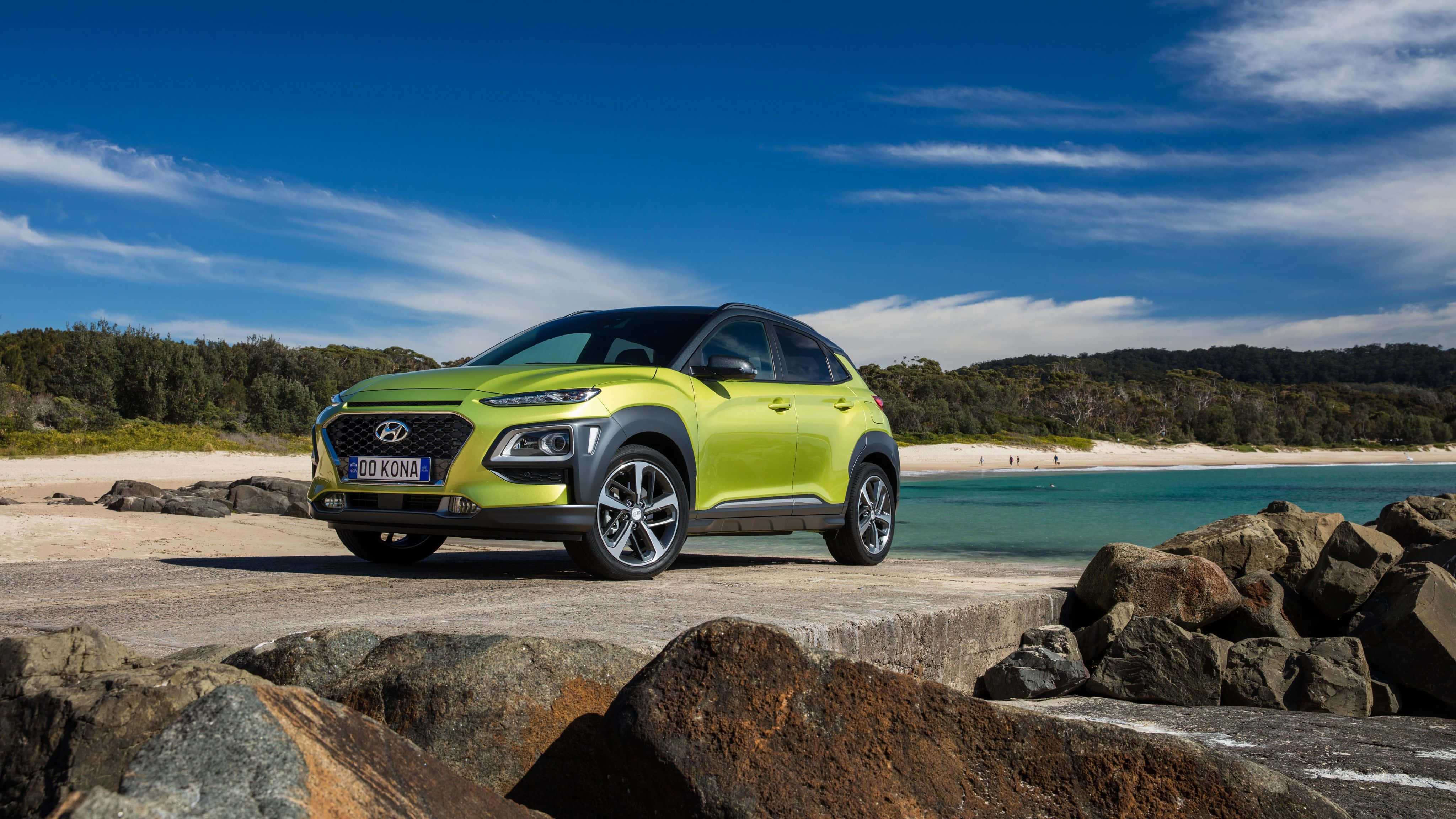 Hyundai Kona 桌布 壁紙 — 一輛充滿活力的萊姆綠色現代Kona SUV停泊在岩石海岸小徑旁，旁邊是碧綠的海水，上方是明亮的藍天。